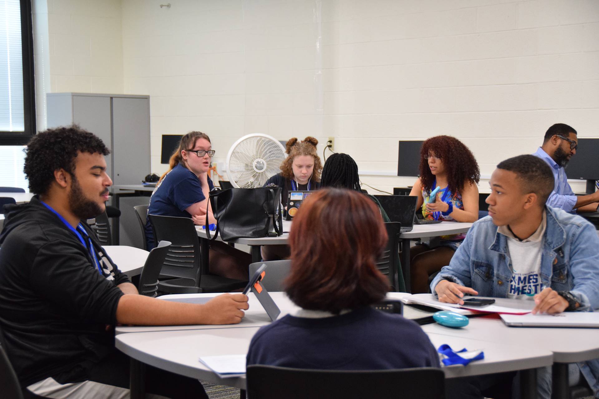 In a well-lit classroom, diverse students work together at round tables. Some are using laptops and tablets. A fan is behind a group of students. A man is working at a computer at a table in the background.