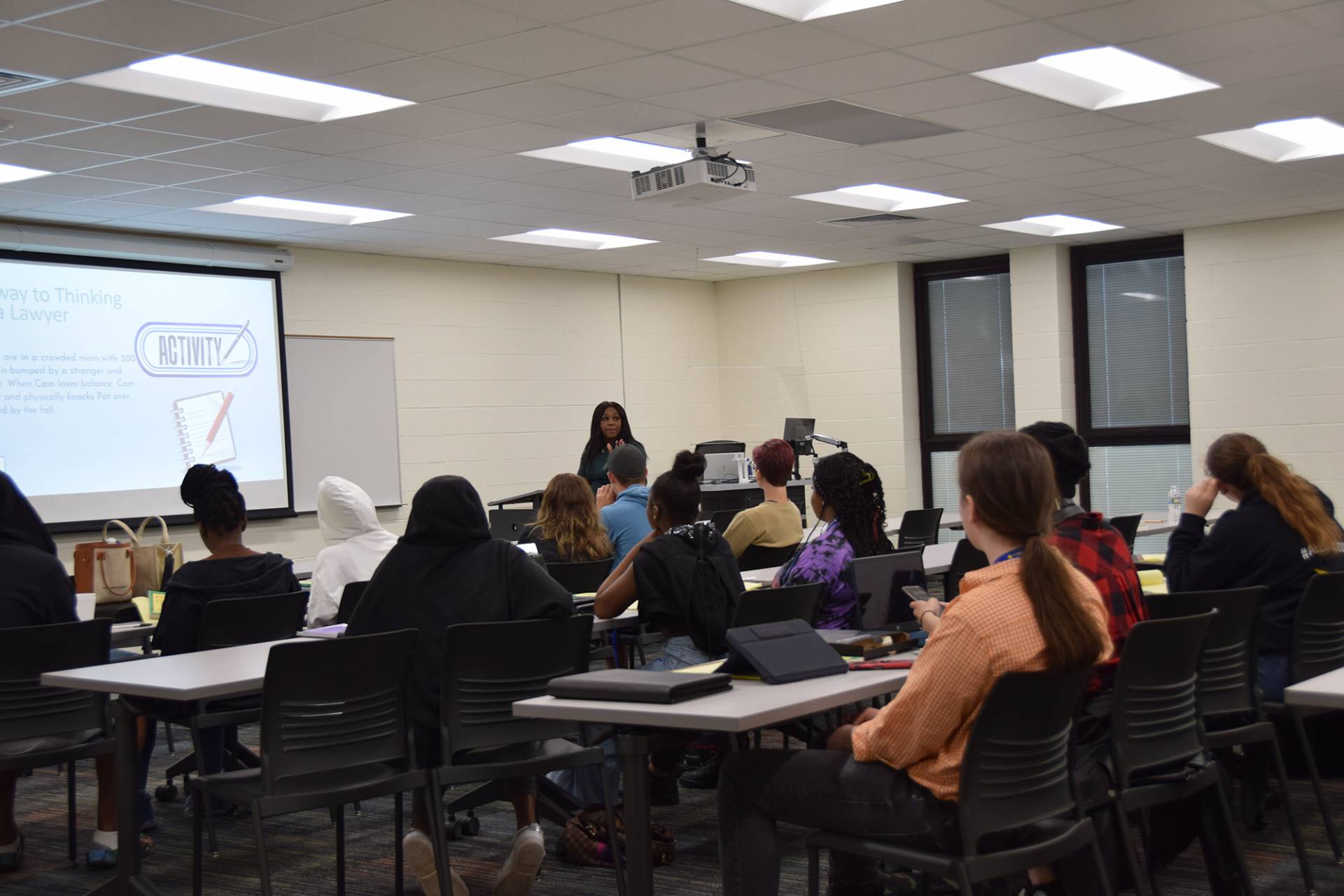 A classroom scene focuses on an female instructor at the front, lecturing to a diverse group of students seated at desks and chairs. A projector screen displays the words "Way to Thinking..." and the word "ACTIVITY" while they are engaged with their laptops or notebooks.