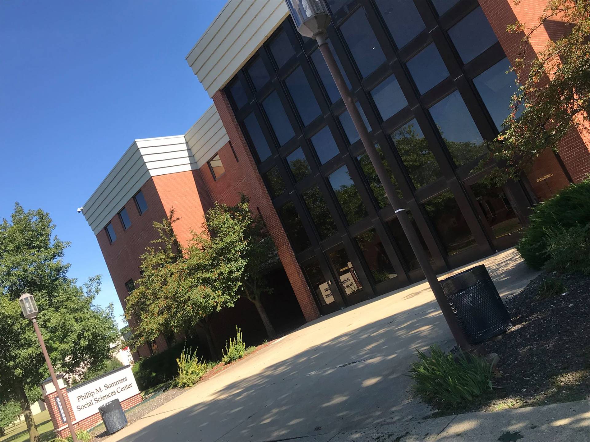 Exterior shot of the Phillip M. Summers Social Sciences Center on a bright day. The building is primarily brick with a distinctive modern design, featuring a large, dark-windowed facade. A sign identifying the building is visible near a black metal trash receptacle. Trees surround the building, and the sky is a clear blue.