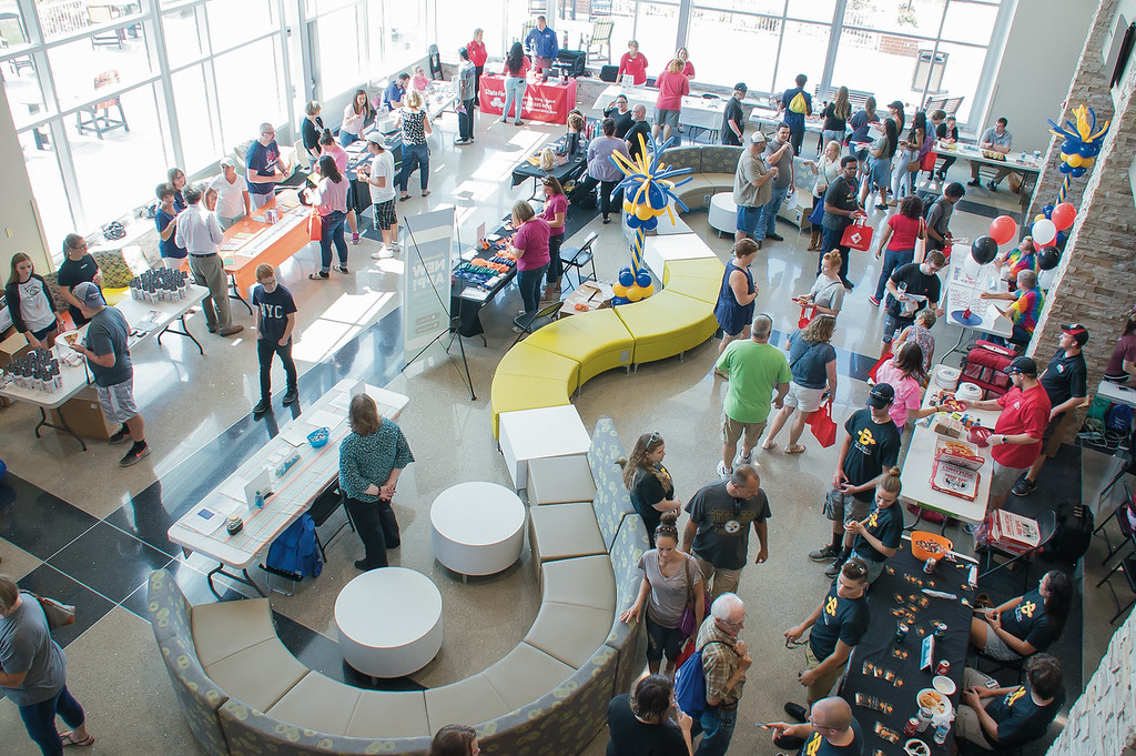 An overhead shot of a crowded event space with many tables and people gathered around them. Natural light floods the space from large windows at the top. Tables are scattered throughout, covered with items and staffed by people engaging with attendees. There are seating areas with curved benches and white cylindrical tables. Many of the attendees are dressed in casual clothes.