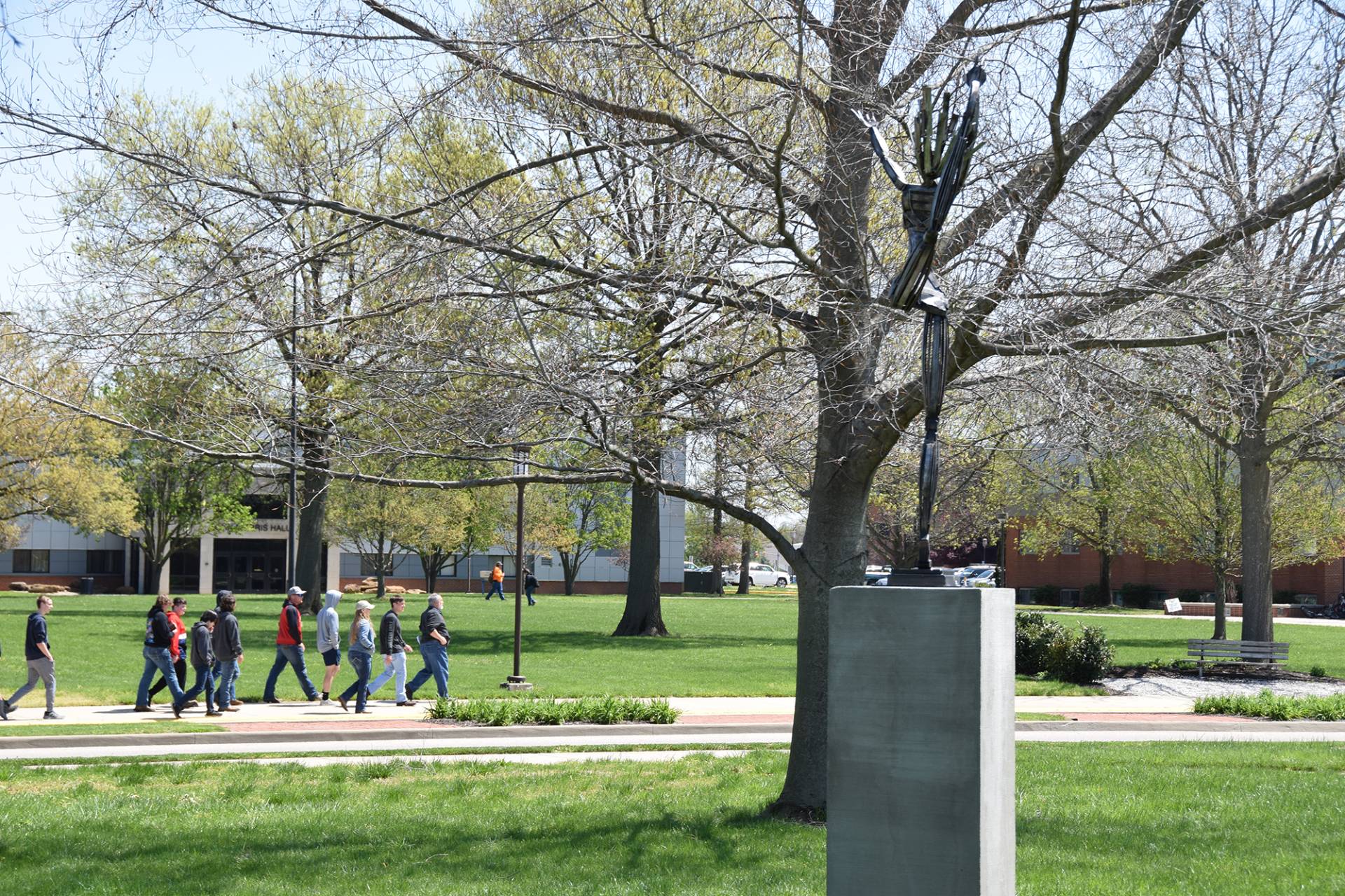 A group of people are walking along a brick pathway on a university campus. They are walking to the left side of the shot and are passing a modern art statue of a person whose arms are raised above their head. The statue is composed of a base and a dark metal statue of a person. Green grass and budding trees populate the background, with buildings visible in the distance under a clear sky.