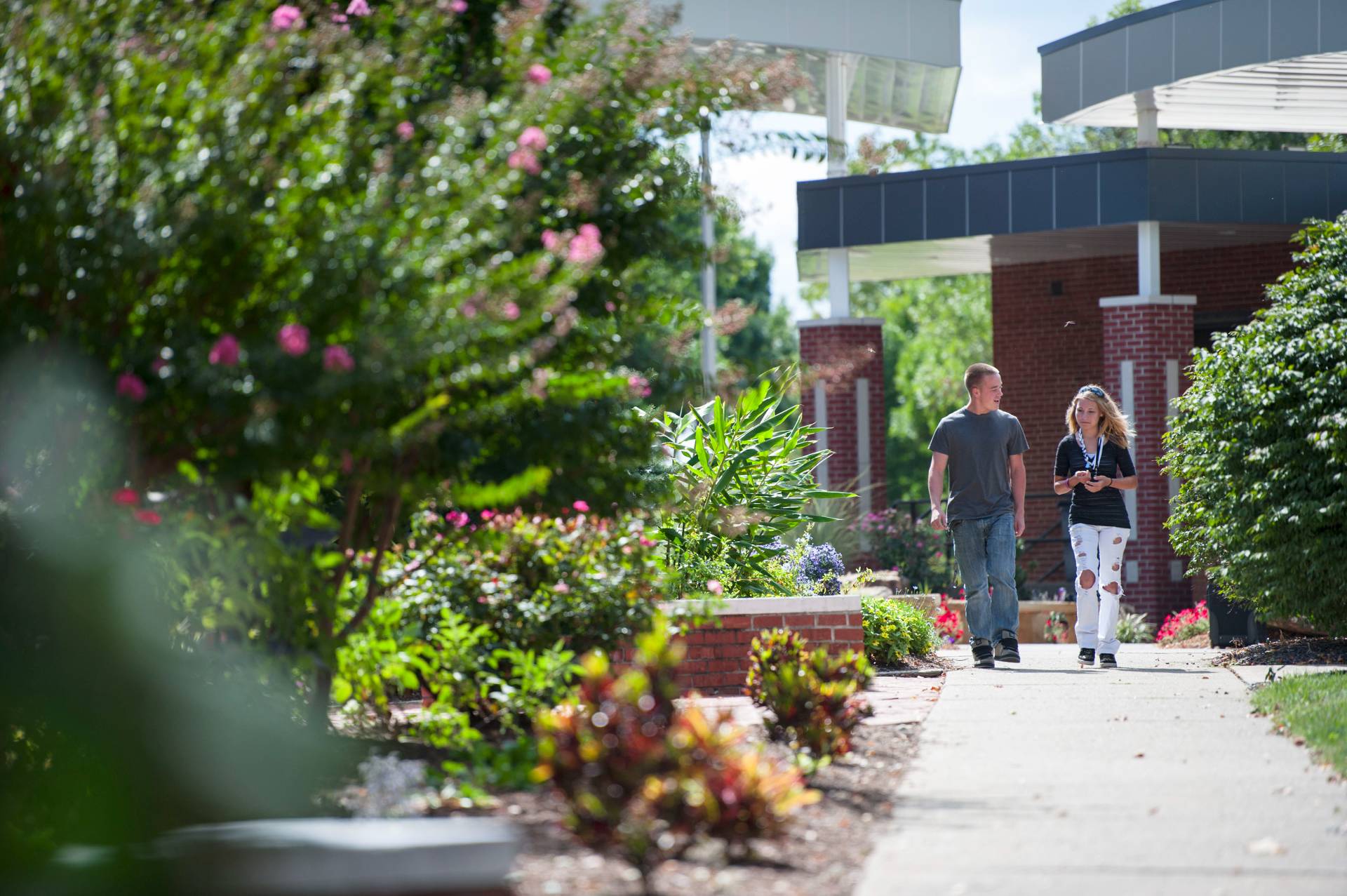 A young man and woman walk along a paved path on a university campus on a bright, sunny day. The woman is wearing ripped jeans and a lanyard. Lush landscaping with flowering trees and bushes frame the path. A building with a modern canopy is in the background.