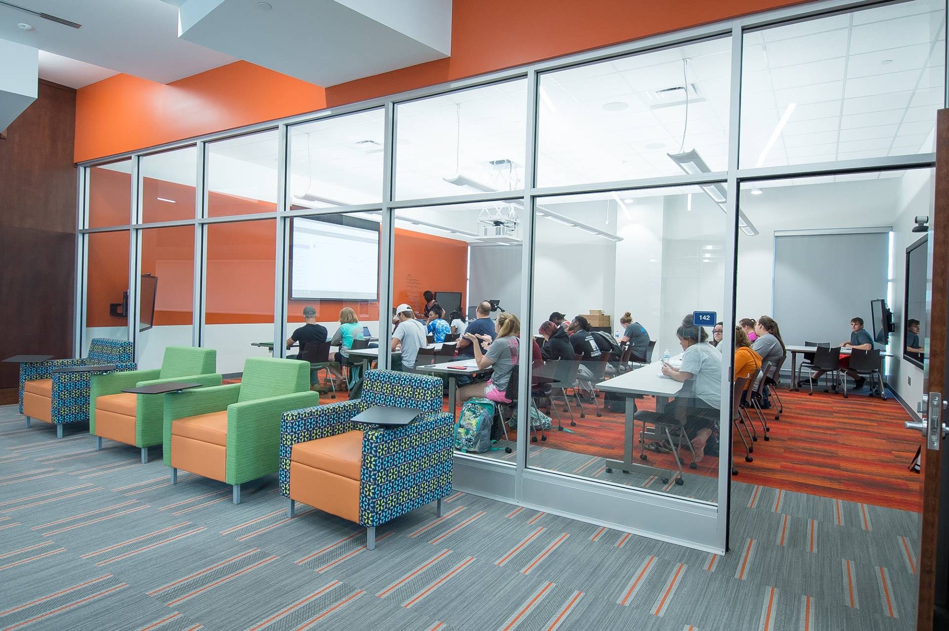 A view through a glass wall into a modern, brightly lit classroom. Students are seated at tables, working individually. A projection screen displays content at the front. In the foreground, outside the classroom, are colorful lounge chairs: three with green arms and one with multi color patterned arms are presented in a row outside the room to be used for other use cases. The room features orange accent walls. The overall impression is of a contemporary and collaborative learning environment.