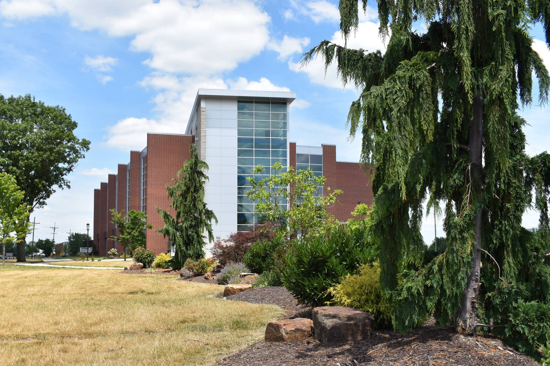Exterior shot of a modern, multi-story brick building with a prominent glass and white paneled facade under a blue, partly cloudy sky. The building is surrounded by a manicured landscape consisting of a light brown lawn, various bushes, trees, and decorative rock features. A large, weeping evergreen tree dominates the right foreground, partially obscuring the building.