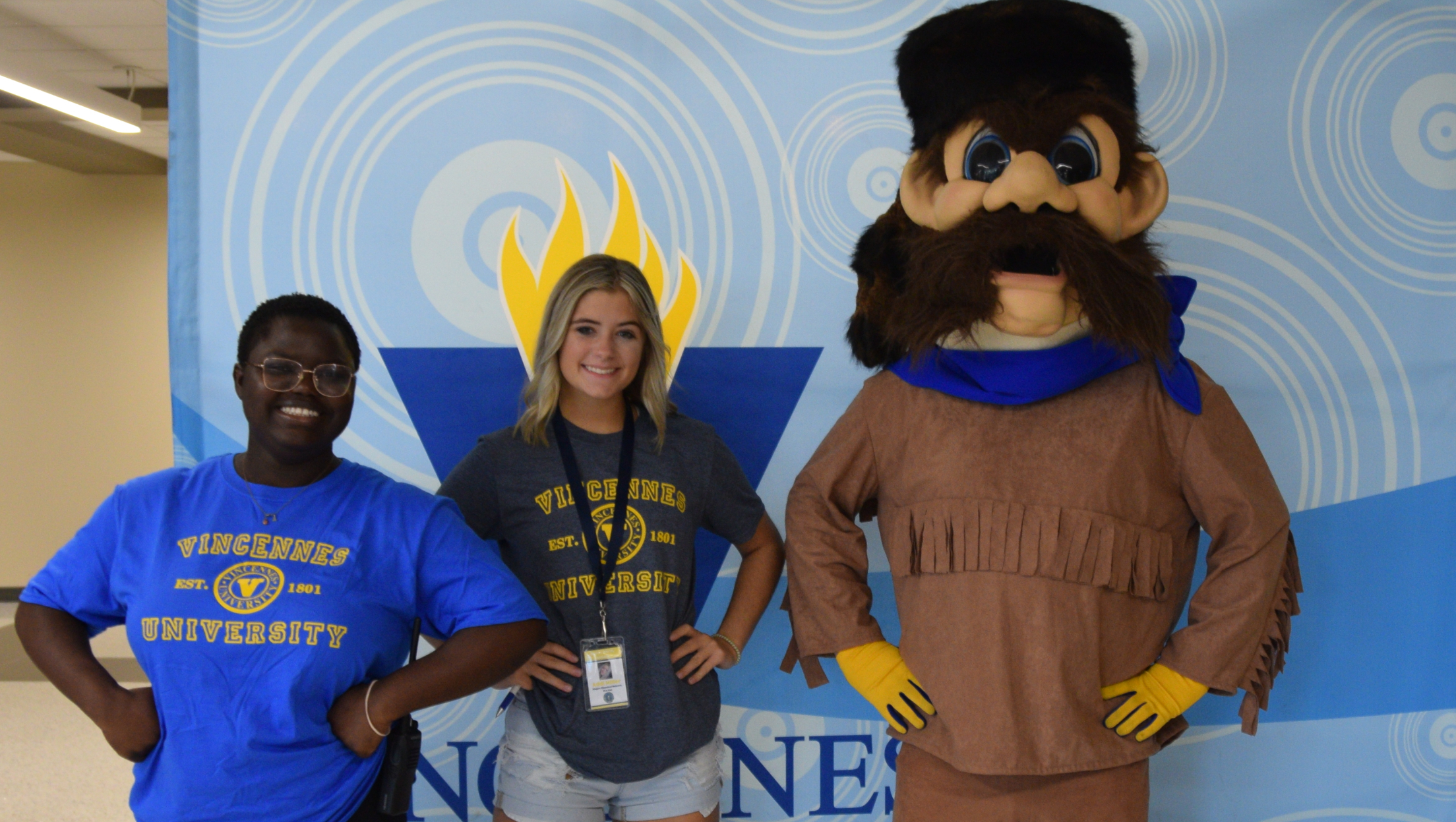 Two smiling female students and the Vincennes University mascot stand together for a photo in front of a backdrop with university branding. The student on the left wears a blue Vincennes University t-shirt and glasses. The student in the center wears a grey Vincennes University t-shirt and has a lanyard with an ID. The mascot is large and furry with a brown costume. The backdrop is blue with concentric circle graphics and the university logo in yellow. Some text is partially visible near the bottom of the backdrop.