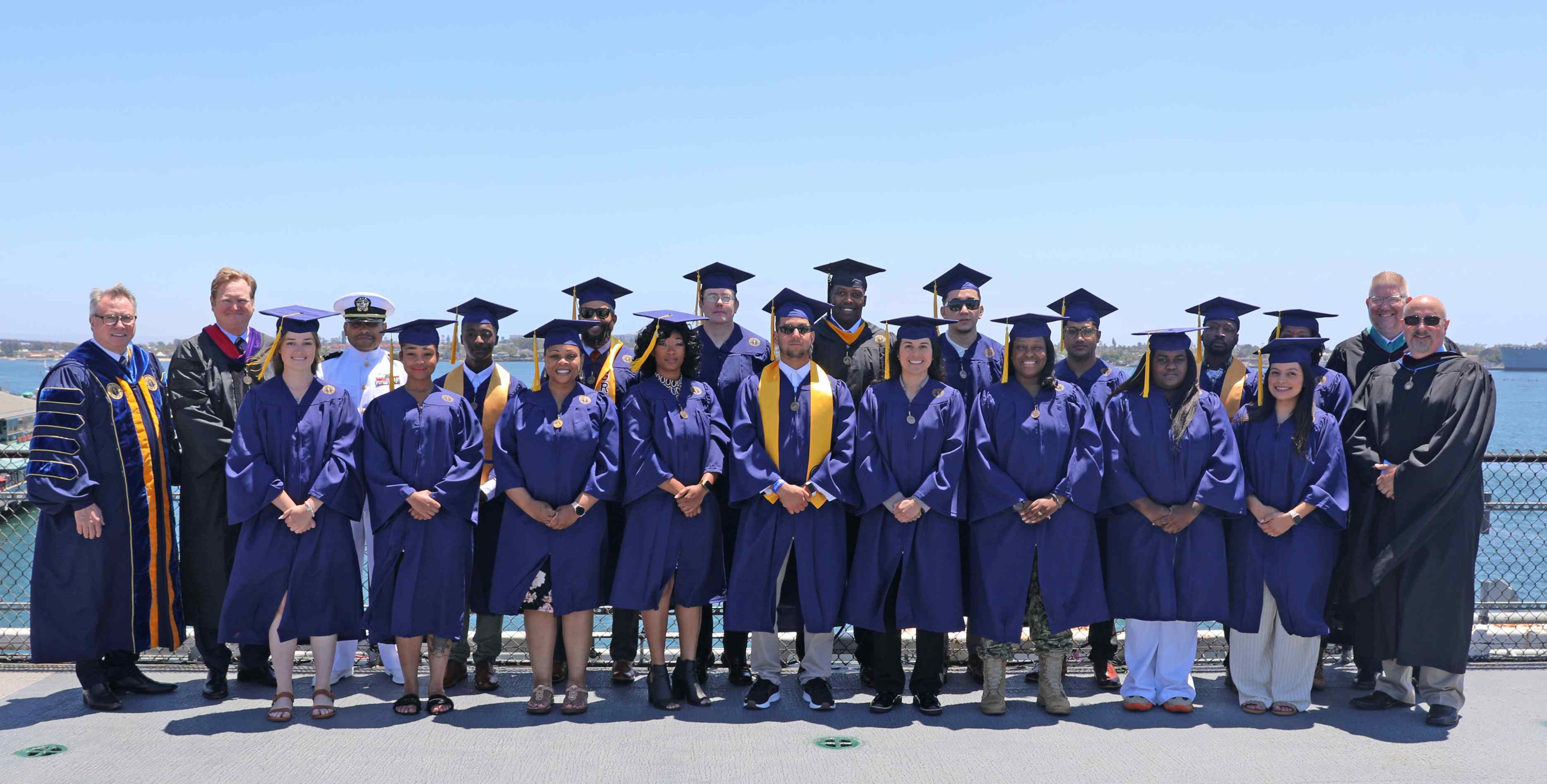 Graduates in blue caps and gowns pose with VU leaders for a group photo outdoors on the deck of a ship during a Vincennes University military commencement.