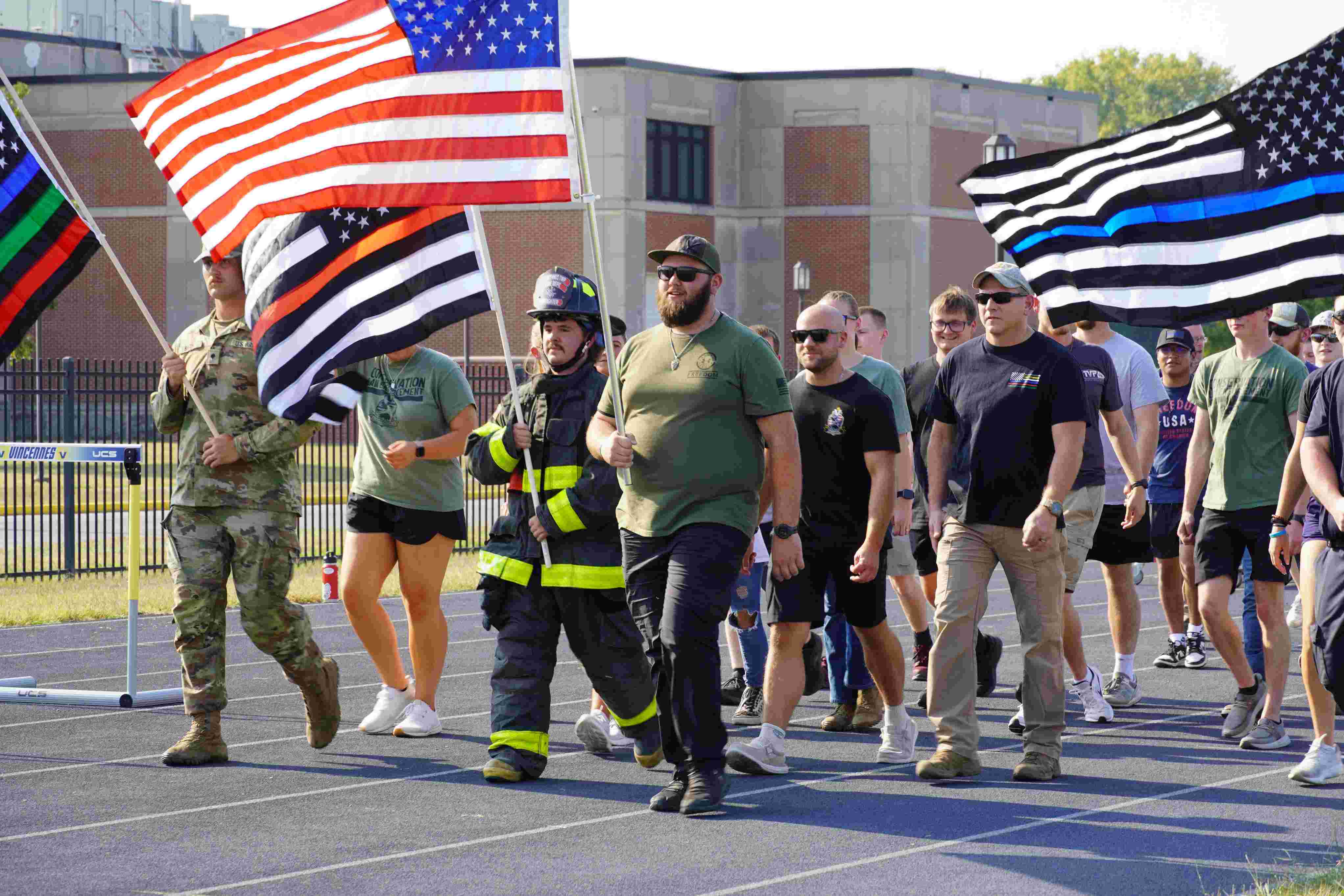 A group of students and faculty walk on an outdoor running track while holding American flags and service honor flags, including a Thin Blue Line flag and a firefighter flag. Participants include a firefighter in turnout gear and military personnel in uniform.