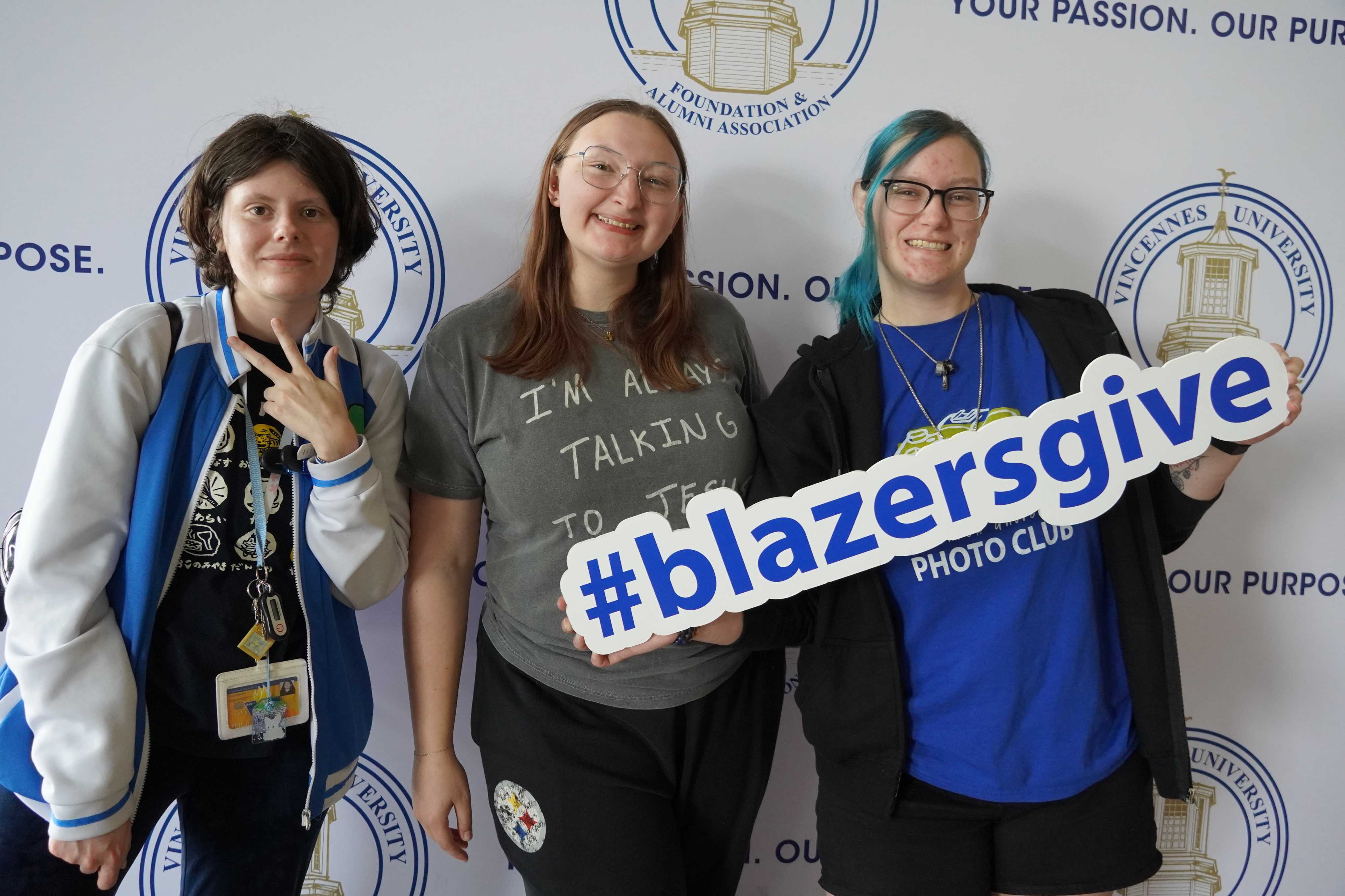 Three students pose in front of a Vincennes University backdrop, giving a peace sign, smiling and holding a large 