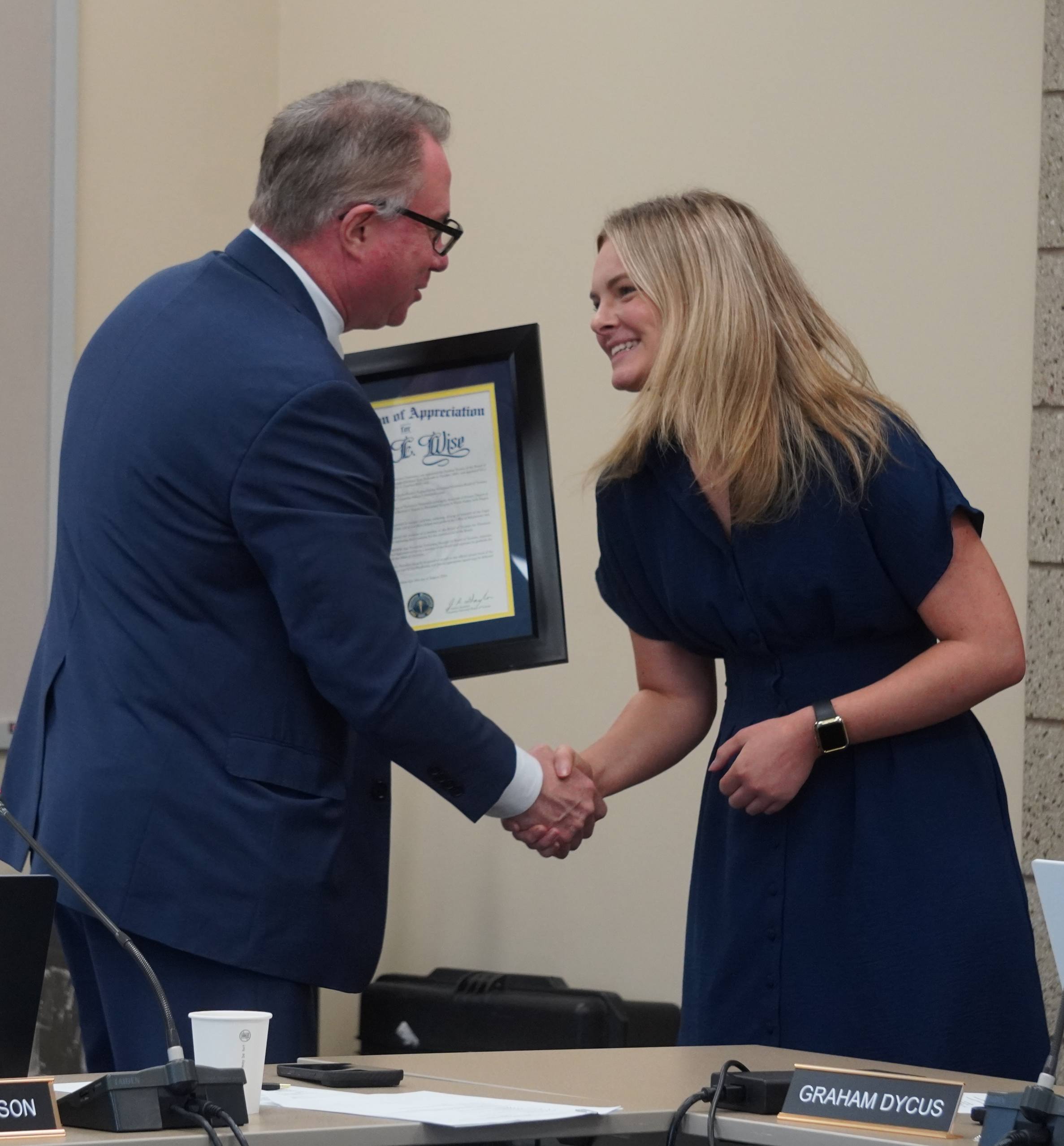 A man in a suit hands a framed certificate of appreciation to a smiling young woman in a dark blue dress as they shake hands.
