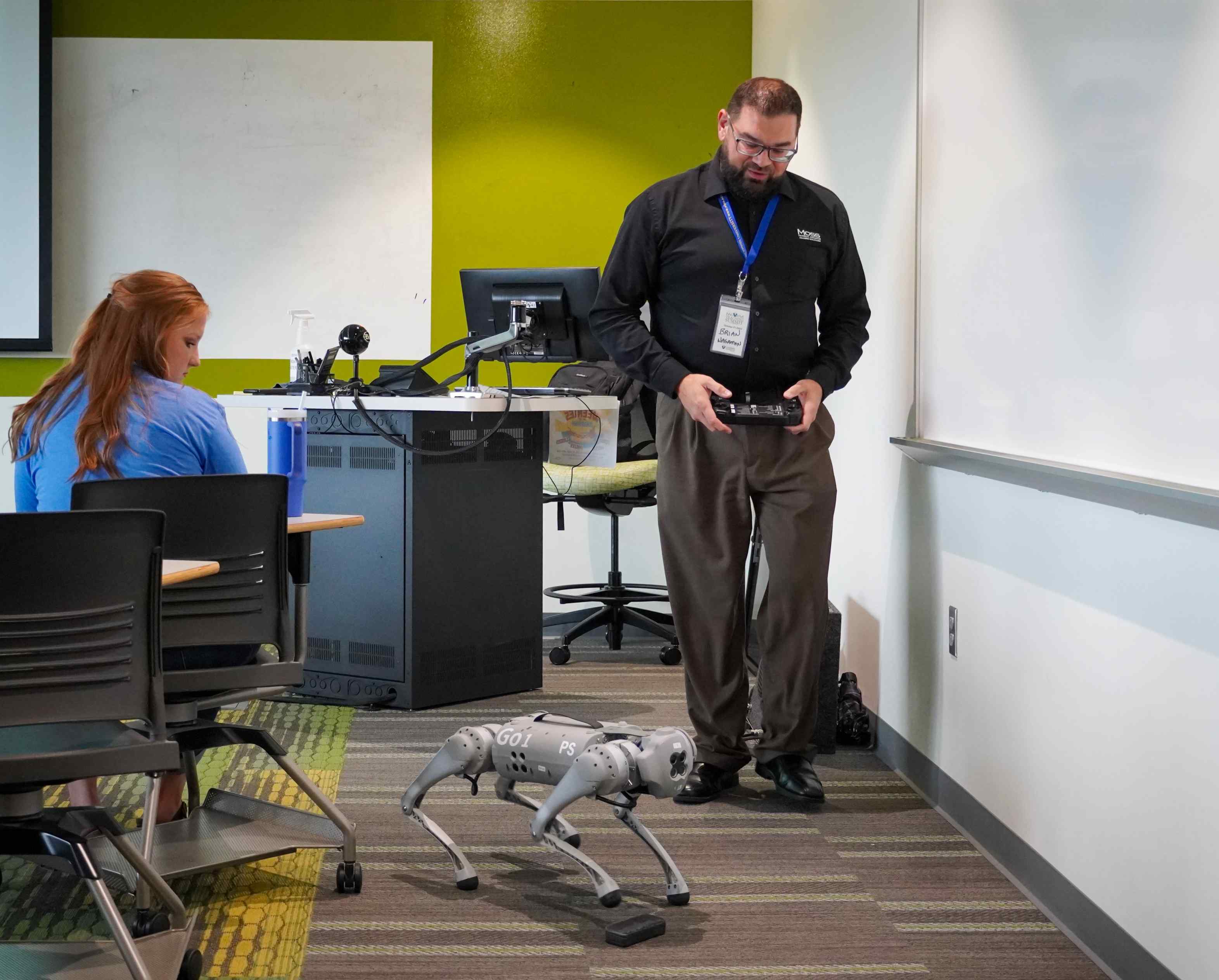 A man holding a remote controller operates a four-legged robotic dog in a classroom while a female sits nearby.