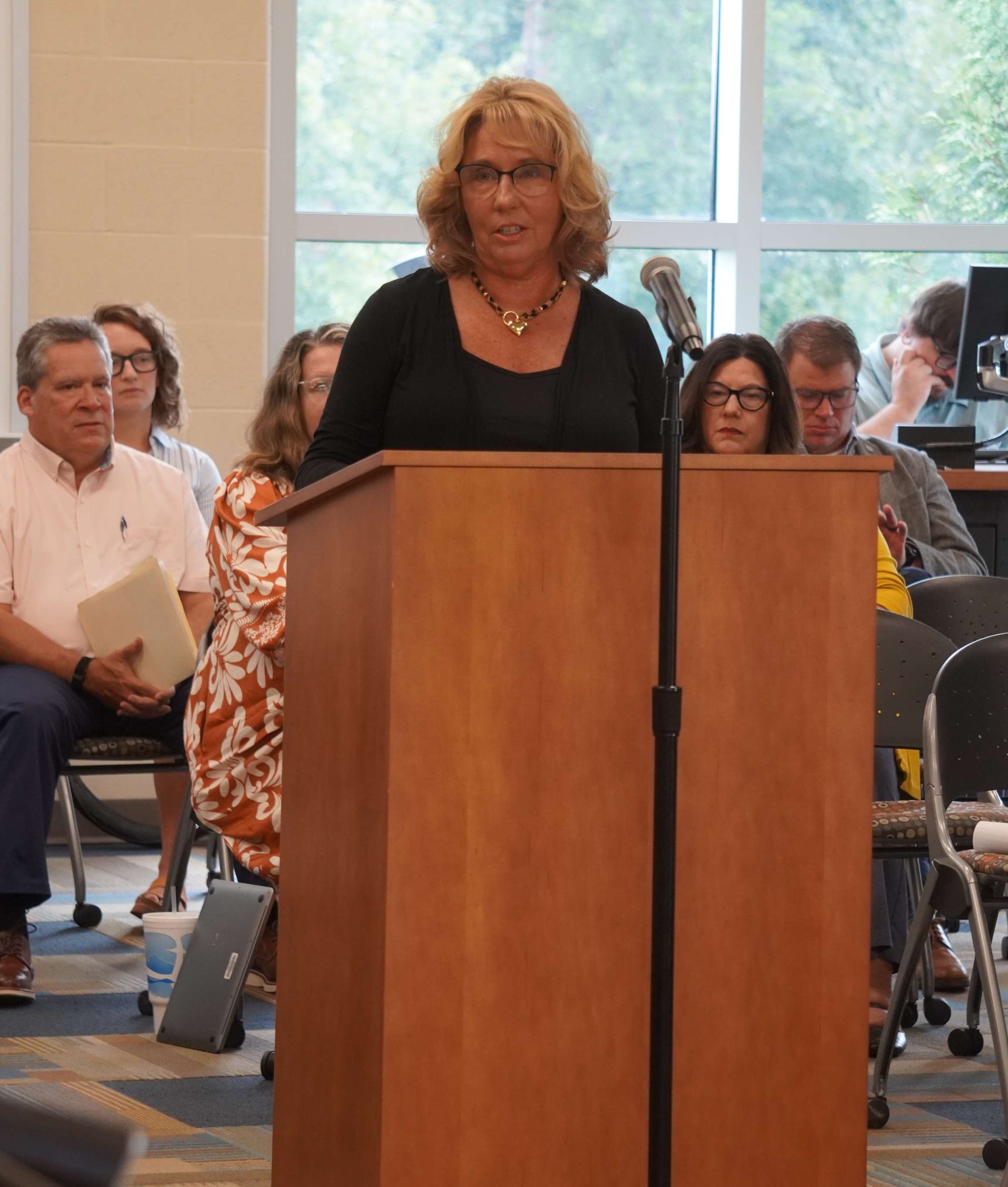 A blonde woman with glasses speaks at a wooden podium with a microphone, addressing an audience seated behind her indoors.