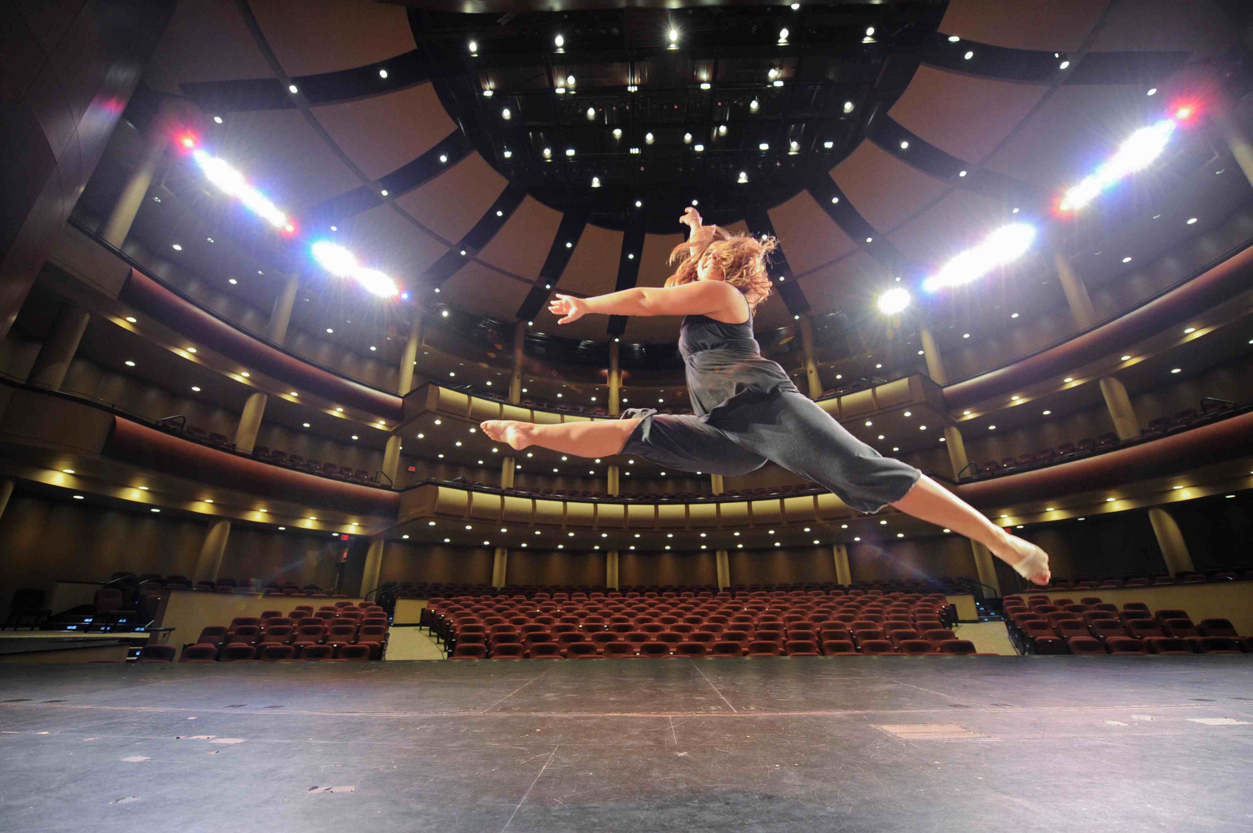 A woman with long hair leaps in the air with arms extended on stage at the Red Skelton Performing Arts Center with theater seating and lights in the background