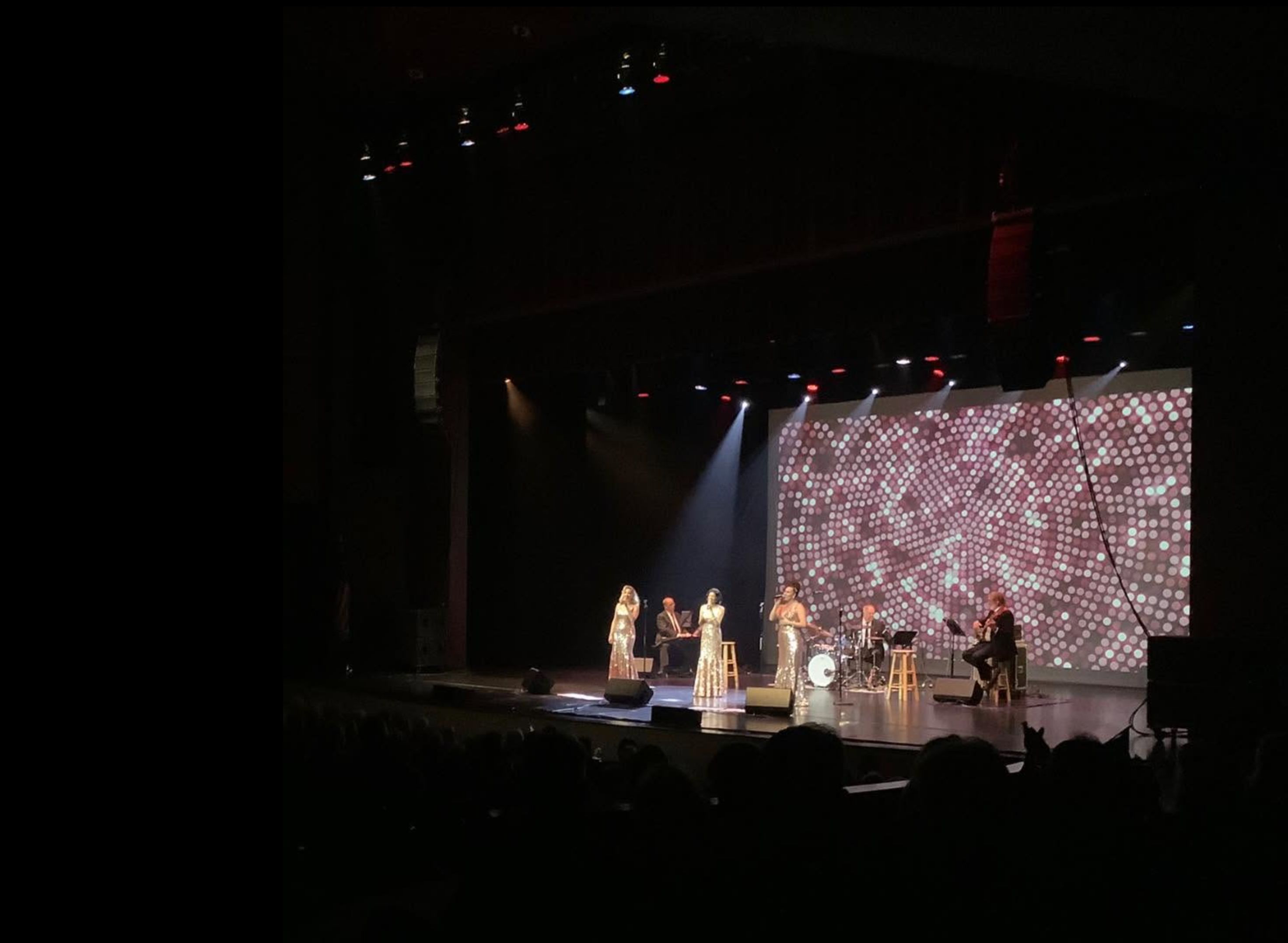 Three singers in sparkling dresses perform on a stage at the Red Skelton Performing Arts Center with a live band, spotlights, and a glowing patterned backdrop screen.