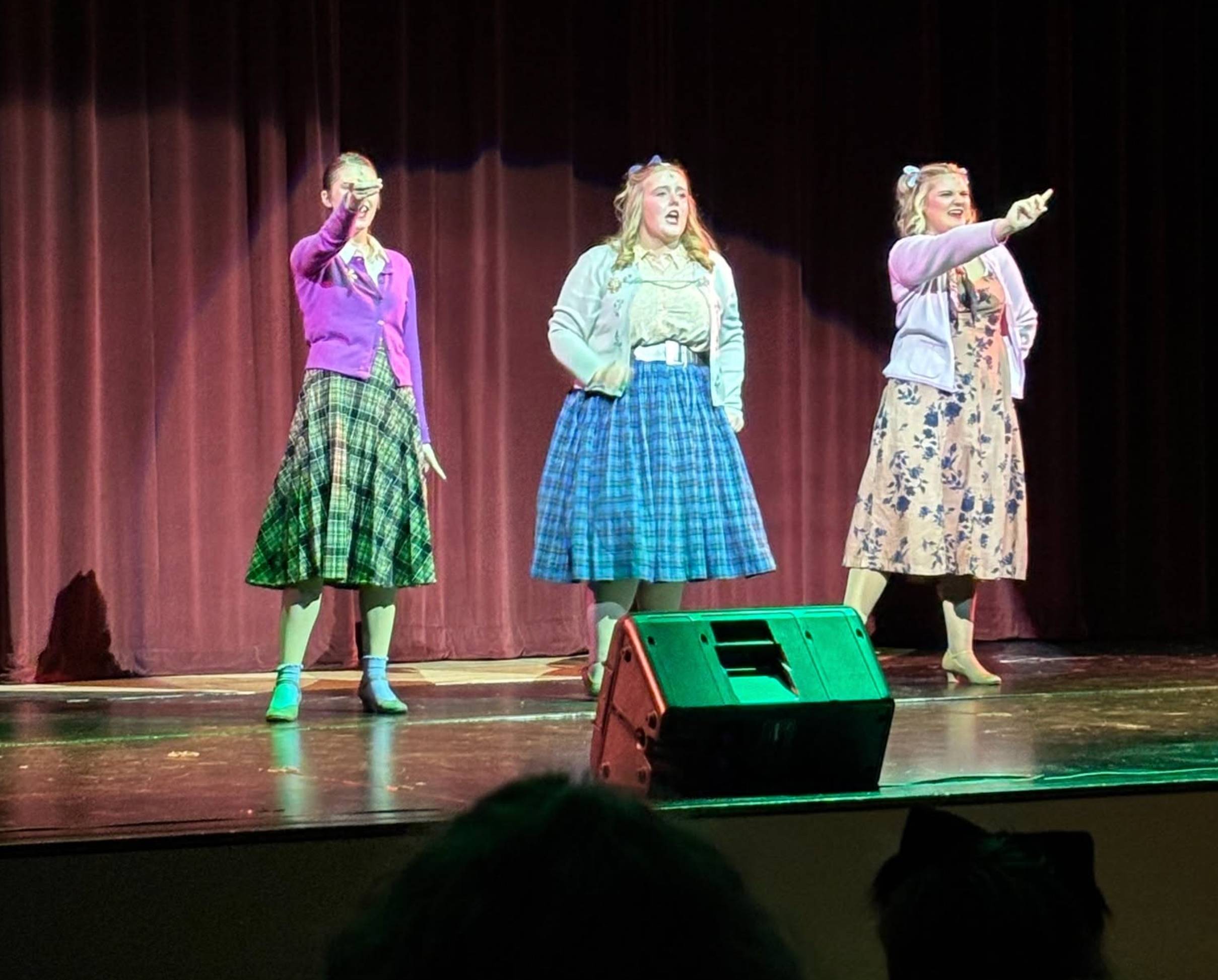 Three student performers in cardigans and skirts sing and gesture dramatic hand poses on stage in front of a red curtain.