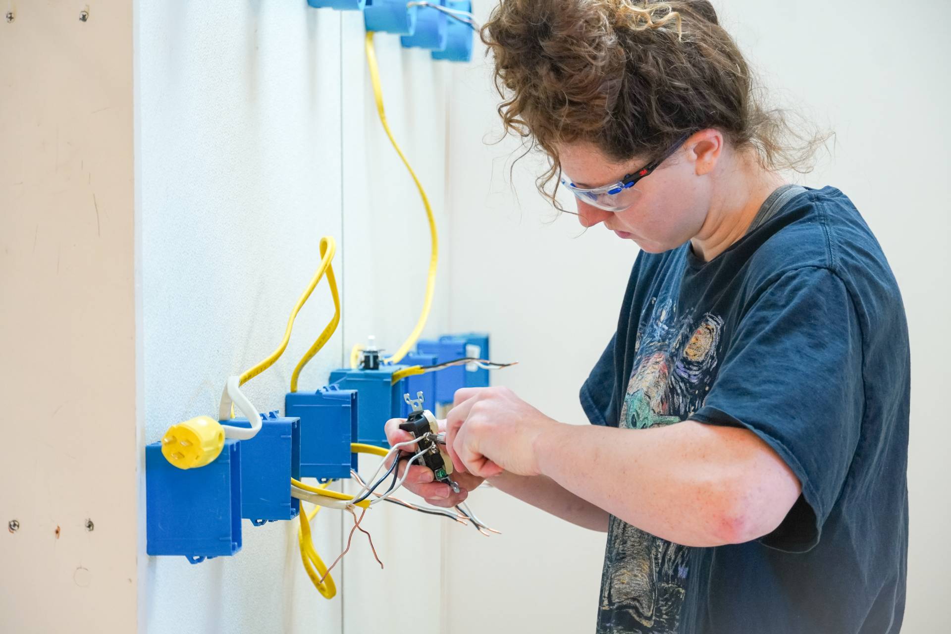 A young woman wearing safety glasses is wiring an electrical outlet on a white wall. She's connecting the wires to a switch, surrounded by several blue electrical boxes connected by yellow cables. The wall has minor scuffs. She is wearing a dark blue t-shirt that features an image of Van Gogh's "Starry Night".