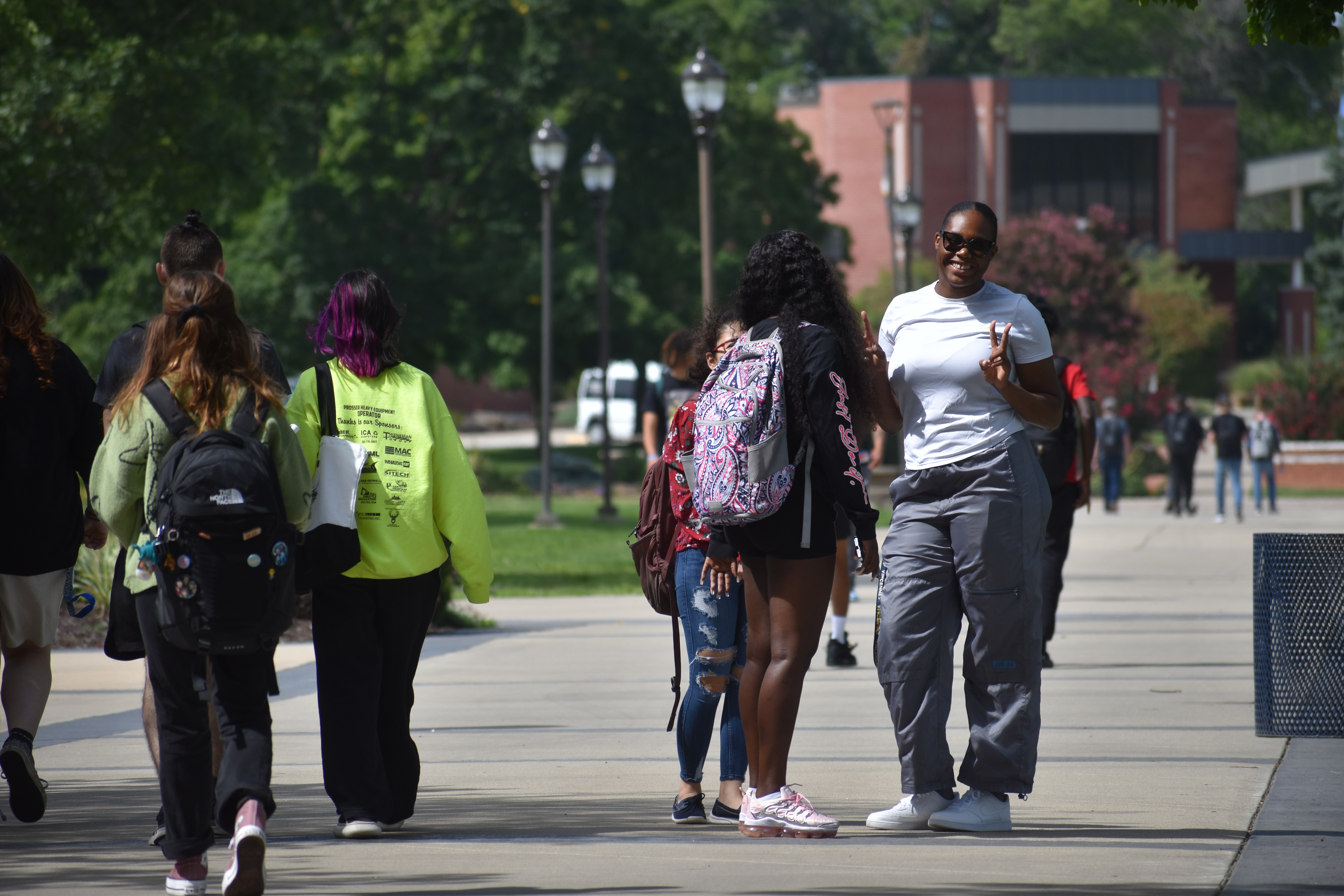 A female student flashes a peace sign on a campus walkway with many students surrounding her.