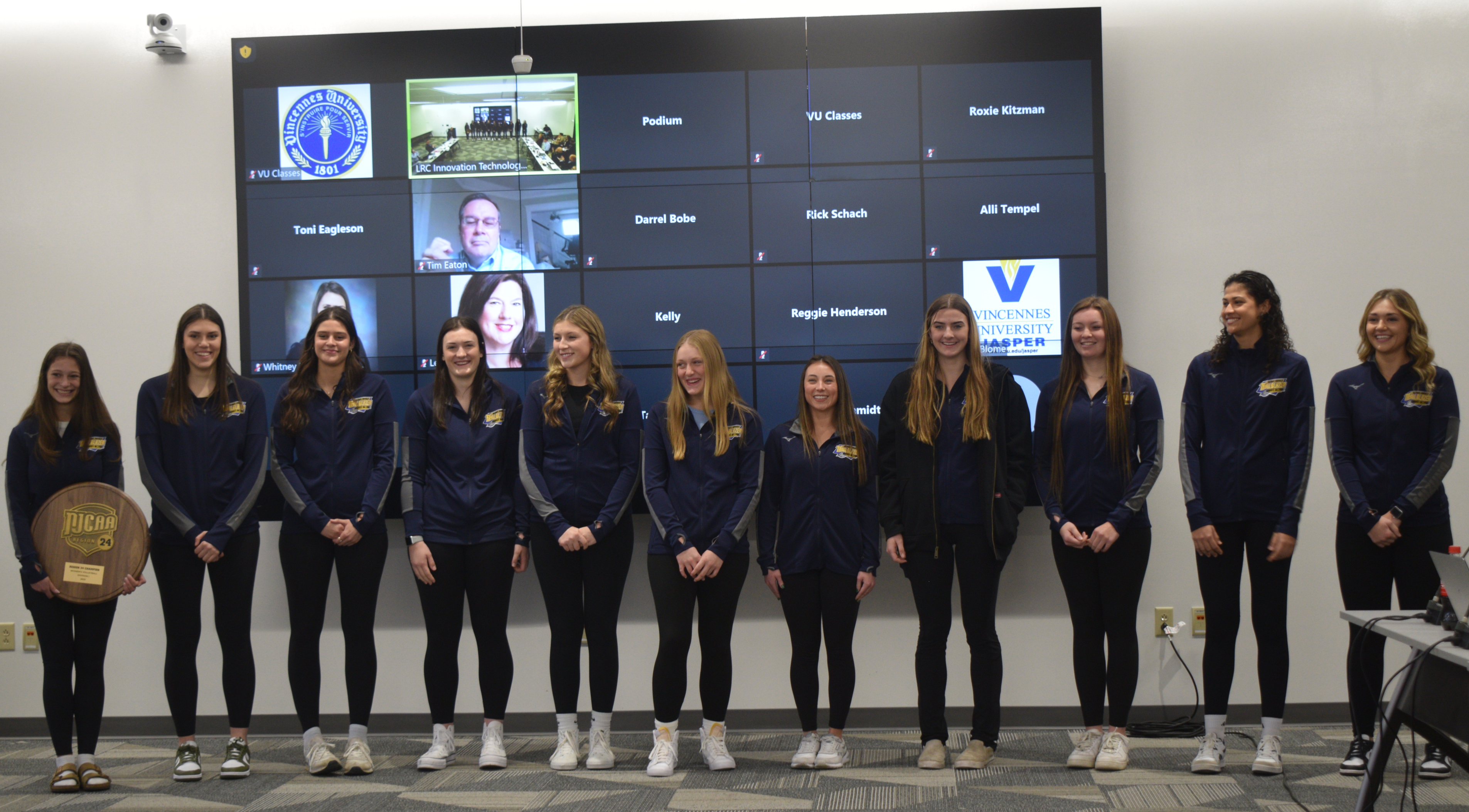 VU volleyball team stands in front of a large screen while one player holds a championship plaque.