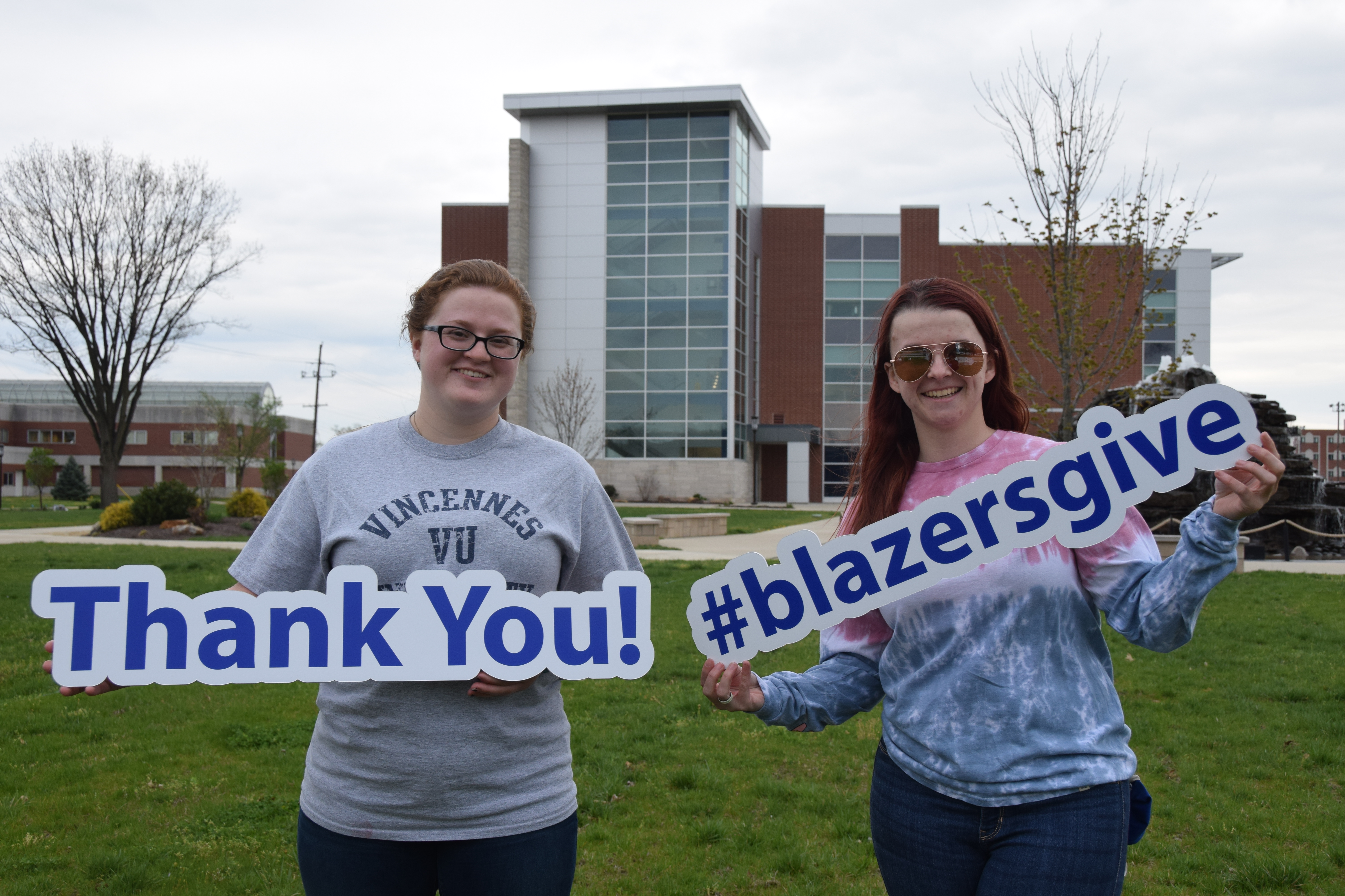 2 female students professor holding Blazers Give and Thank you hashtag signs in front of Updike Fountain