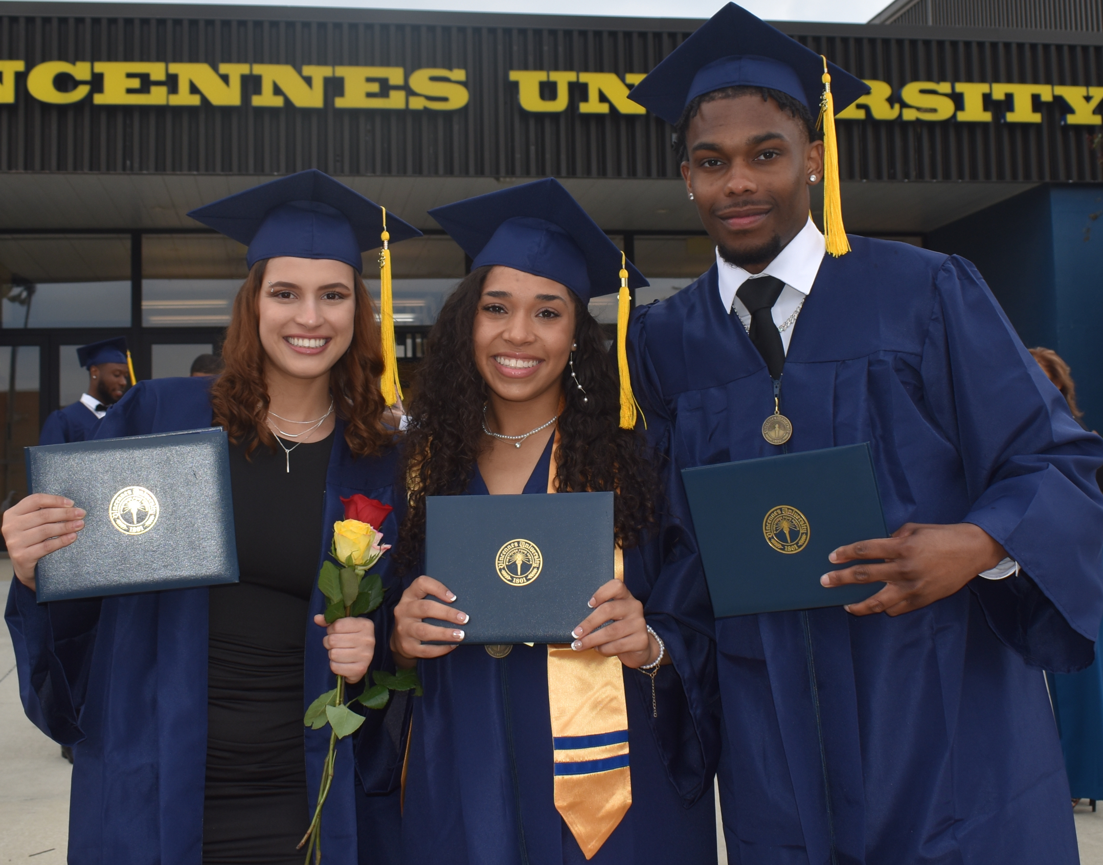 2 female and 1 male VU students wearing graduation caps and gown holding their degrees with a Vincennes University sign behind them.