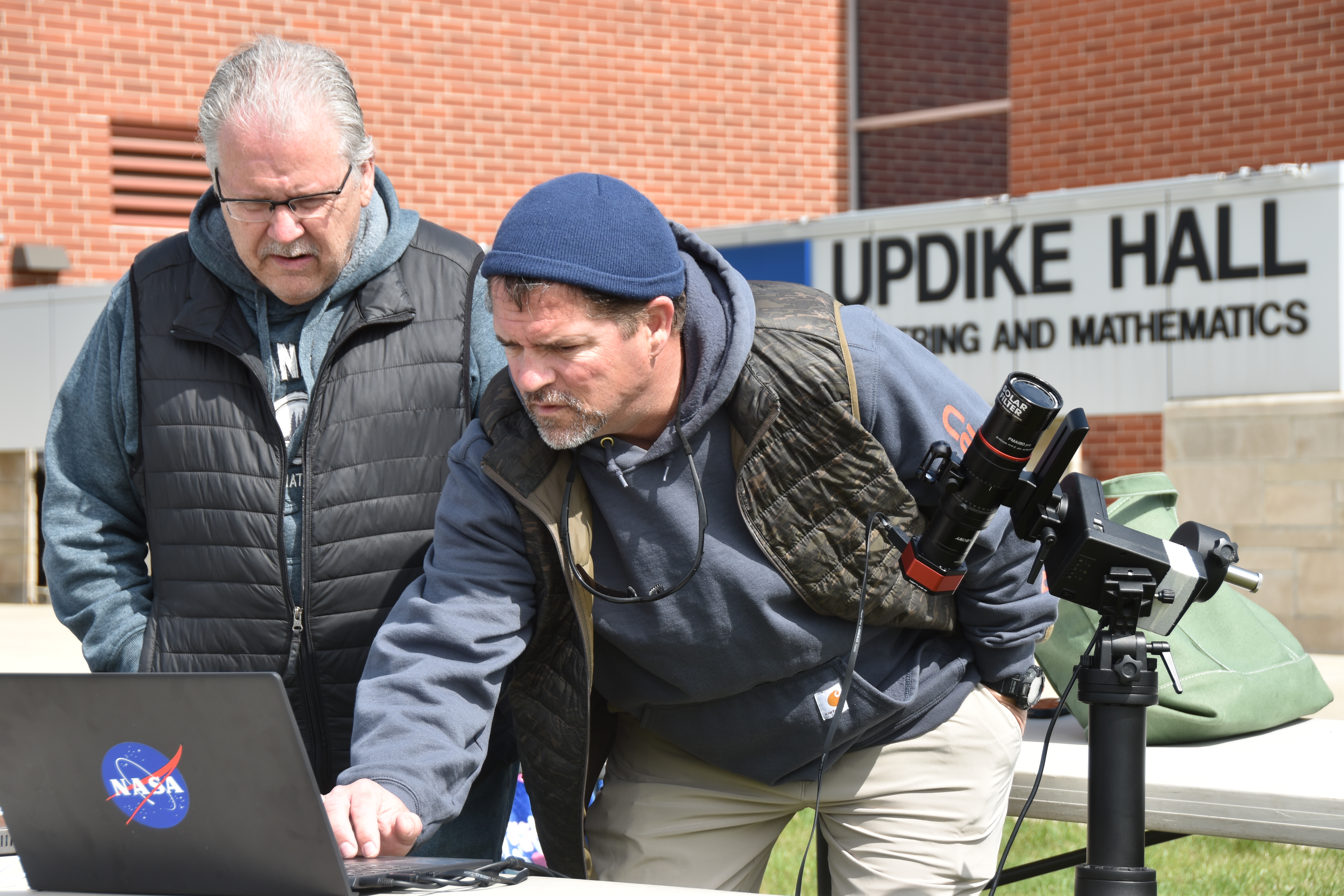 Two males looking at a laptop screen outside Updike Hall. The laptop has a NASA sticker on it. 