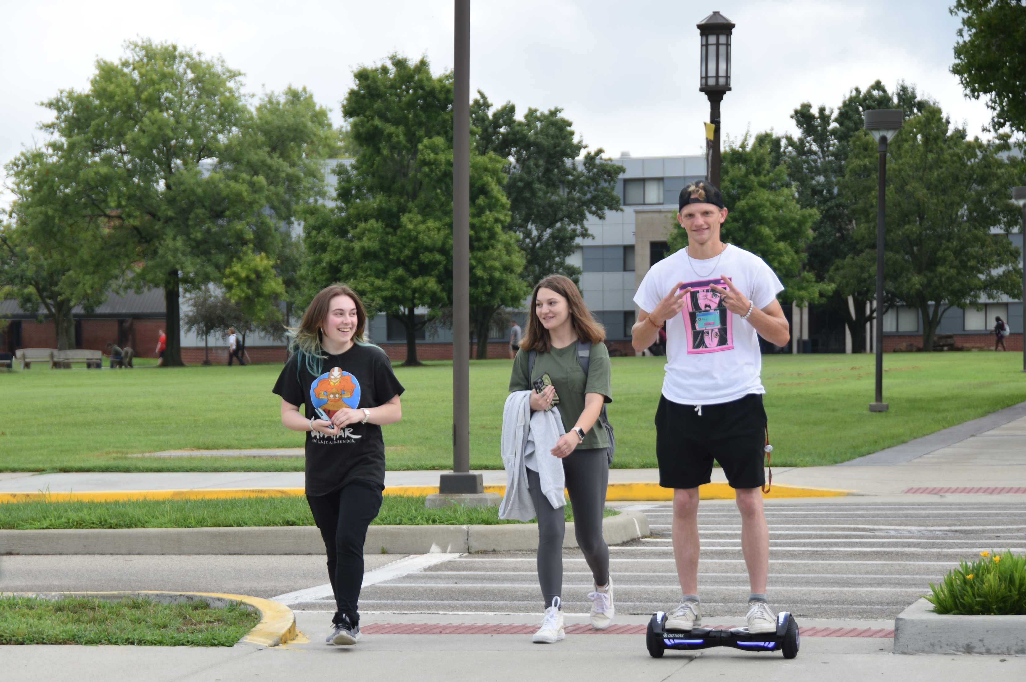 A male student flashing a peace sign and riding a hoverboard on campus with 2 female students next to him.