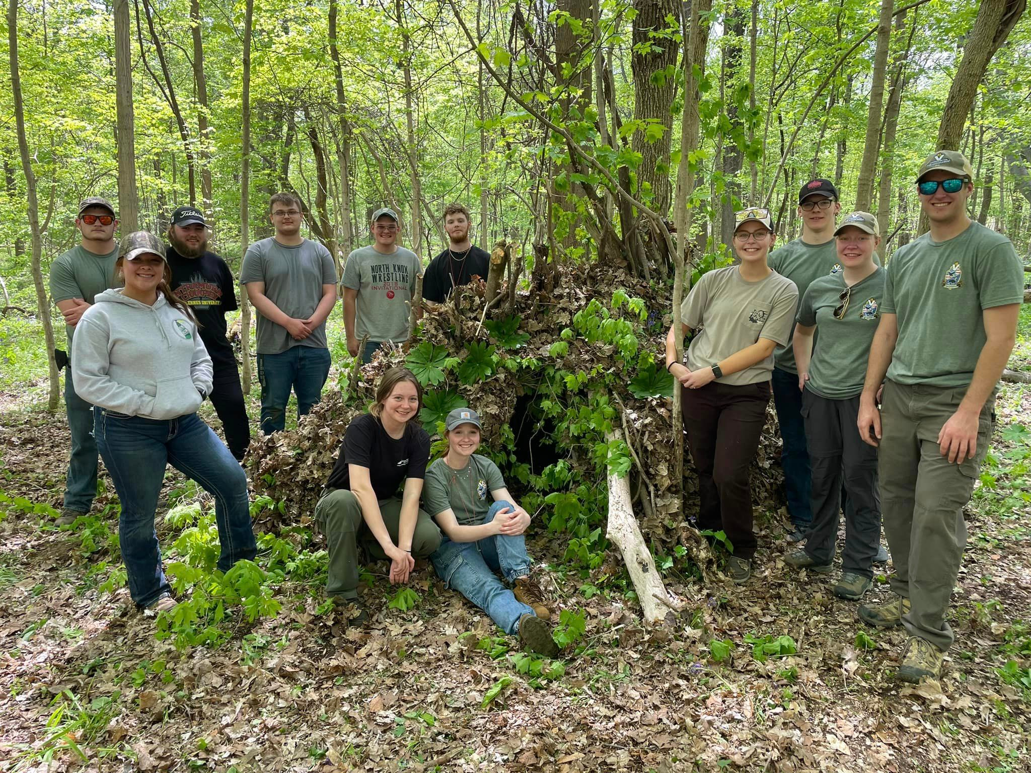 A group of Conservation Law Enforcement students stand in front of a shelter they built with natural materials