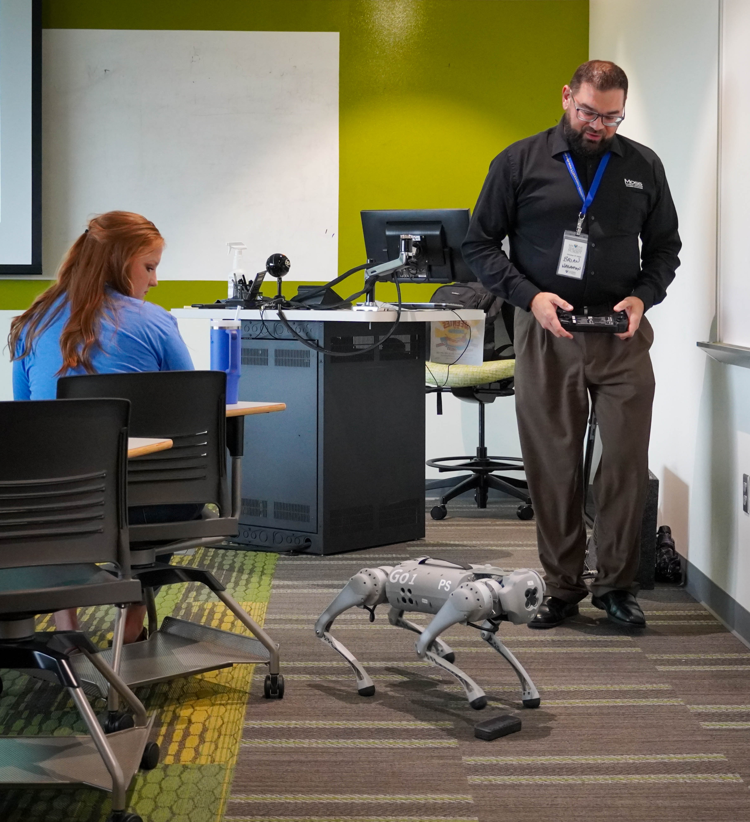 A male operates a robot dog in a VU classroom as a woman looks on.