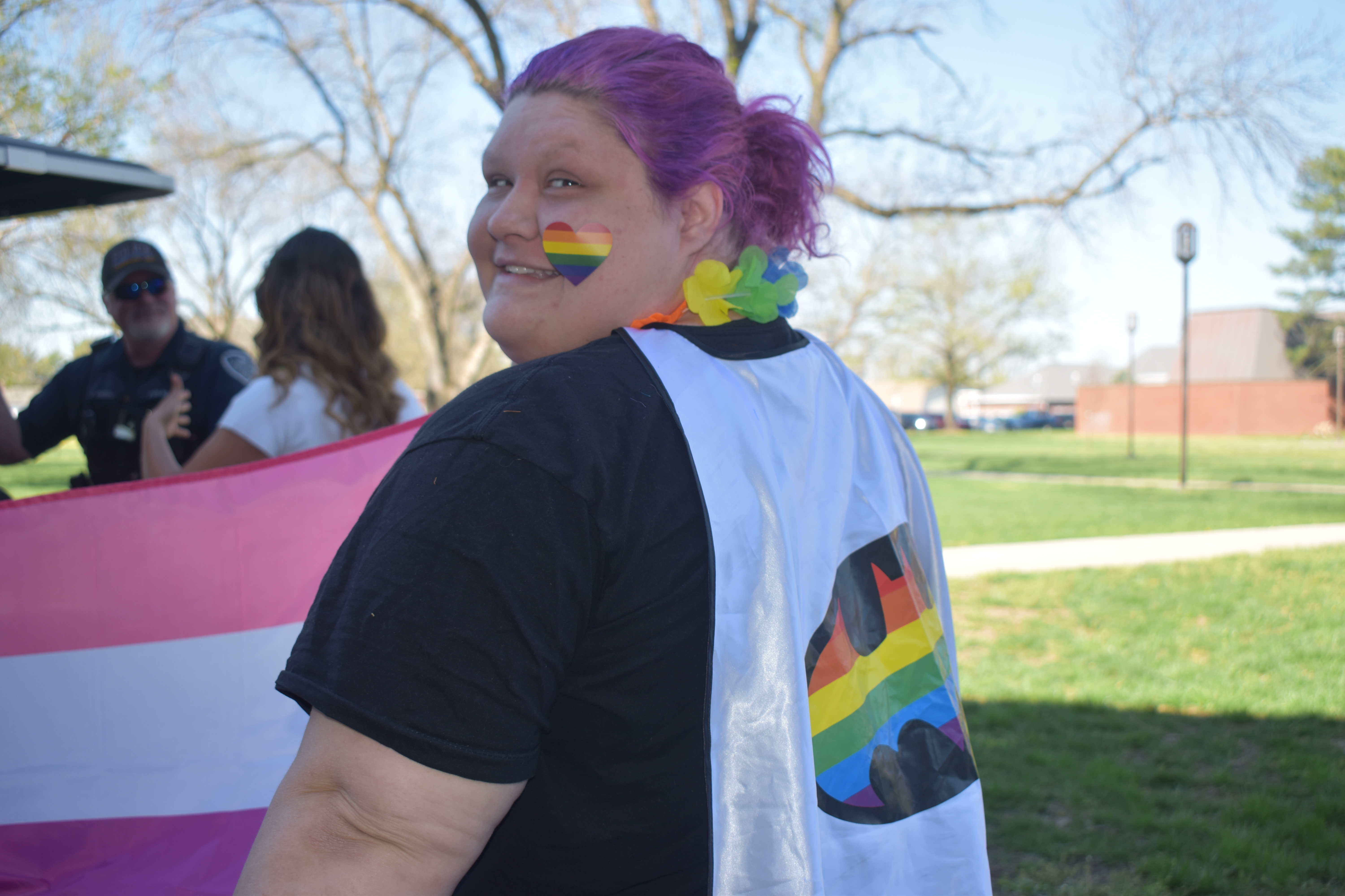 A student wearing a rainbow heart sticker on cheek and a cape with a rainbow logo on the back