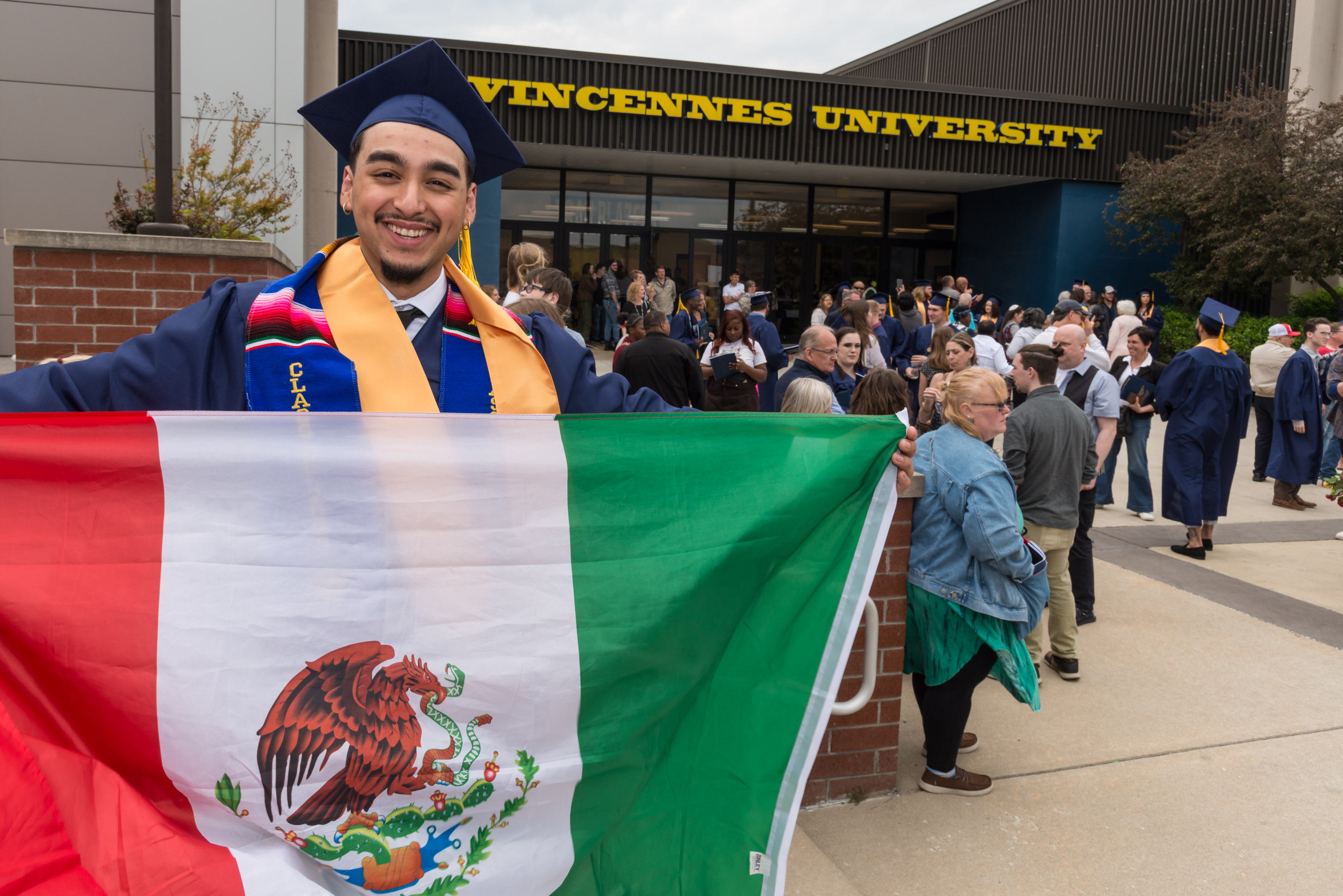A male VU graduate wearing a cap and gown holds a Mexican flag outside of P.E. Complex