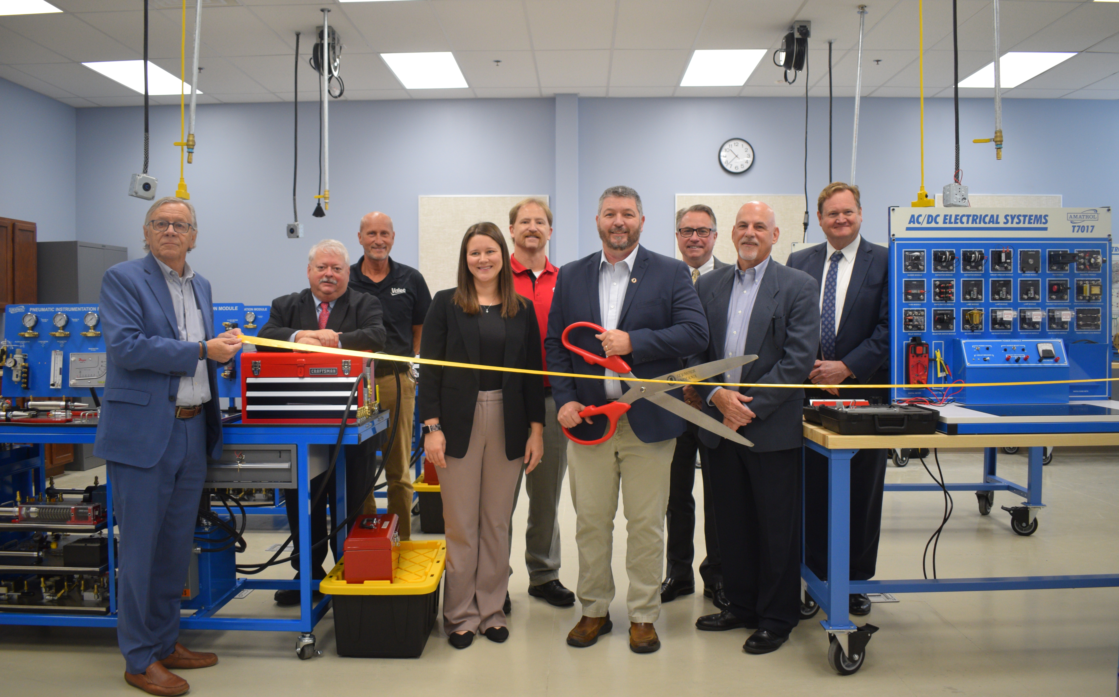 City, county, state and VU officials pose for a photograph as they cut a ribbon on the VU industrial maintenance lab. A man is holding a large set of scissors while another holds the yellow ribbon. The group is surrounded by training equipment.