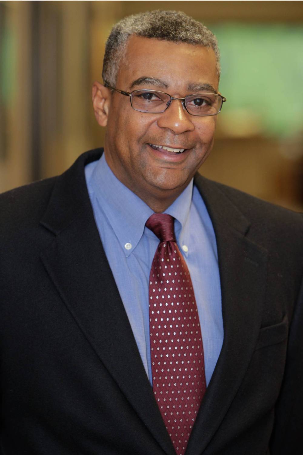 Reginald Henderson, with short gray hair, is pictured from the chest up, wearing glasses, a navy blue suit jacket, a light blue button-down shirt, and a maroon tie with a small diamond pattern. They are looking directly at the viewer with a warm expression.