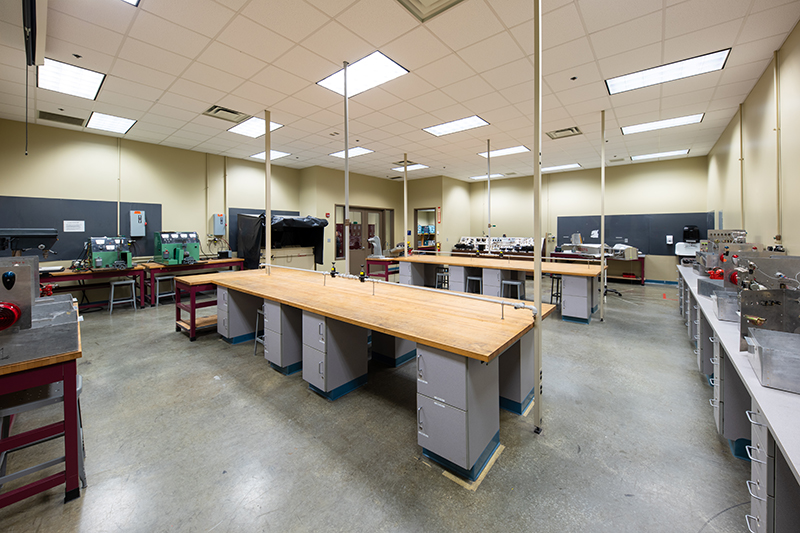 An engineering laboratory classroom features workbenches, fixed equipment, and storage. The room is well-lit with multiple overhead fluorescent lights. There are several workbenches with wooden tops. Vertical poles run from floor to ceiling and are seemingly anchored mid-workbench. The back and side walls are lined with benches holding more complex equipment, alongside blackboards.