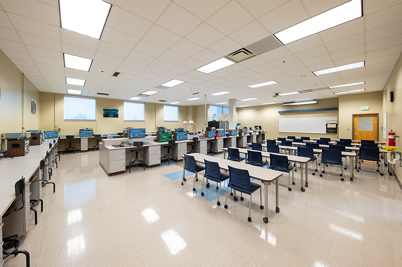 A brightly lit electronics lab classroom. The room is divided into two sections: student desks with blue chairs arranged in rows for lectures, and individual workstations with electronic equipment and swivel chairs along the walls. There is a whiteboard, an exit door, and safety equipment visible, against the upper right wall. The classroom is clean and well-organized.