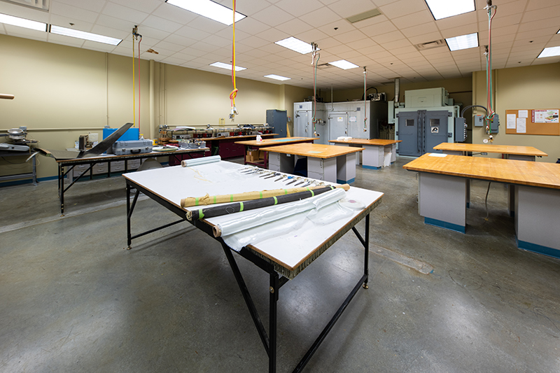A well-lit composites laboratory featuring several workstations and specialized equipment for materials testing and fabrication. In the foreground, a large table holds rolls of composite fabrics and associated tools, suggesting ongoing or recently completed work. Additional work tables are arranged throughout the room, with testing machinery and larger equipment pieces visible in the background.