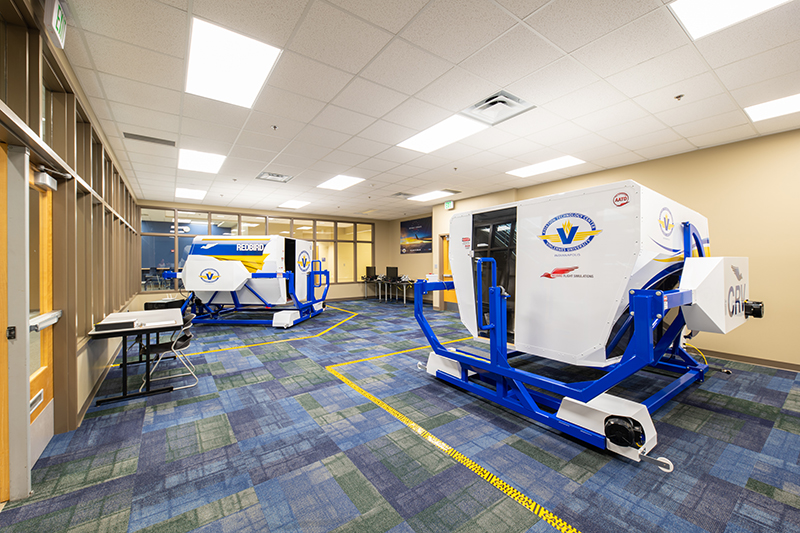 An interior view of a flight simulator training room, featuring two simulators. The simulators are white with blue trim and the logos of the aviation training center. The room has a blue and green patterned carpet, and is well-lit with drop ceiling lights. Yellow safety lines denote the operational areas of the simulators.