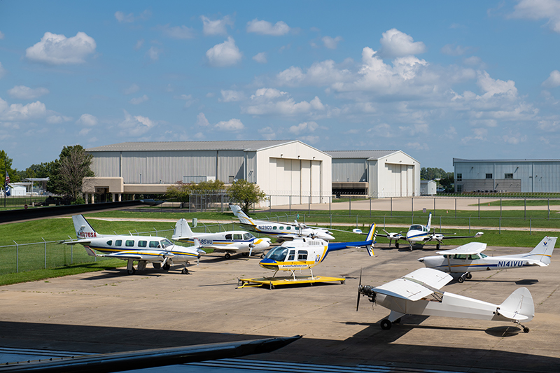 An aerial view captures an active airfield on a bright day. A collection of several parked airplanes, including a yellow and blue helicopter, sits atop the paved tarmac in the foreground. Beyond the aircraft, multiple large, light-colored hangars stand against the backdrop of a partly cloudy blue sky. Fencing and landscaping outline the perimeter of the airfield.