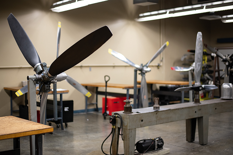 A workshop scene featuring aircraft propeller maintenance or repair. Several propellers are mounted on stands, with various tools and worktables scattered throughout the space. The propellers are silver and some have yellow tips. The room is brightly lit with fluorescent lights, and the overall atmosphere suggests a practical, hands-on learning or repair environment.