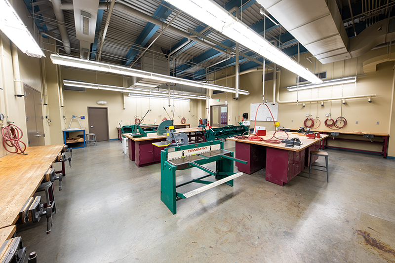 A metalworking classroom features various tools and equipment on workbenches. The equipment includes a shear press, a punch press, and a cut-off saw. The space is well-lit with industrial lighting.