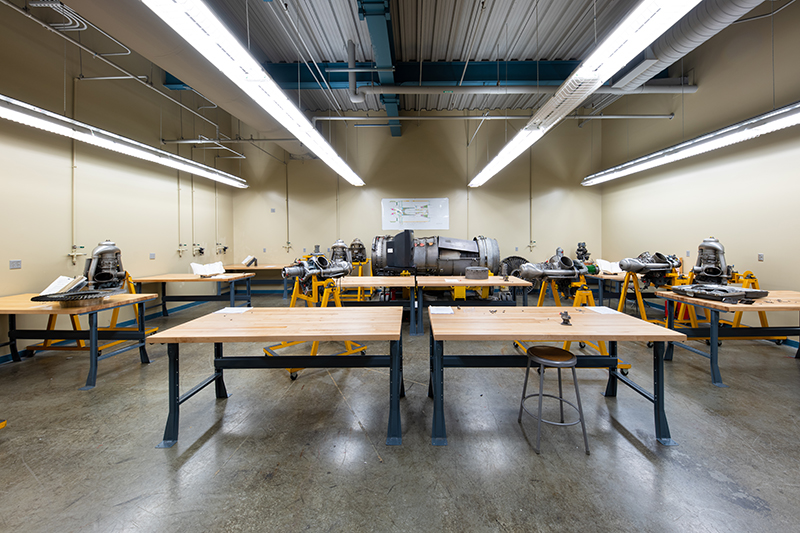An aviation maintenance training lab features several light wood workbenches with various aircraft engine components positioned on top. Some components are mounted on yellow stands. A full engine is in the center, with a schematic hung on the wall behind it. Fluorescent lights illuminate the room in rows.