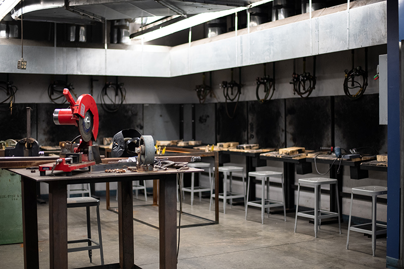 Interior view of a metalworking or welding lab. In the foreground, a metal worktable features a red chop saw and a grey grinding wheel. Additional worktables with stools line the back of the room beneath overhead ventilation hoods and mounted welding equipment. Dark marks are on the wall from previous welding work.