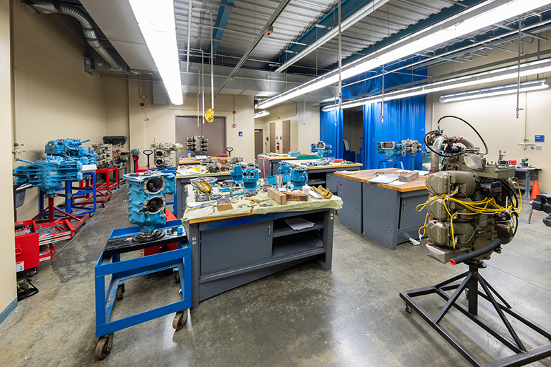 A bright, well-lit mechanical engineering lab features multiple workstations with engines, various tools, and equipment. Some engines are aqua; others are industrial green. Blue workbenches and rolling carts are visible. Overhead fluorescent lights illuminate the space, which is organized and functional with an industrial aesthetic.