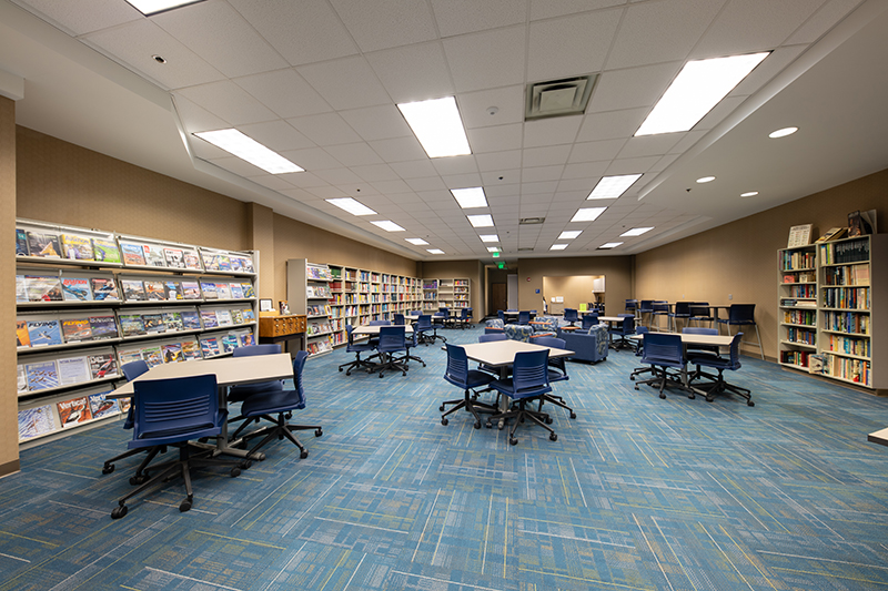 A well-lit library reading room features rows of bookshelves lining the walls, filled with books and magazines. Several square tables with blue chairs are arranged across a blue patterned carpet. In the background are some blue sofas in a lounge area. The room is brightly illuminated by recessed ceiling lights.