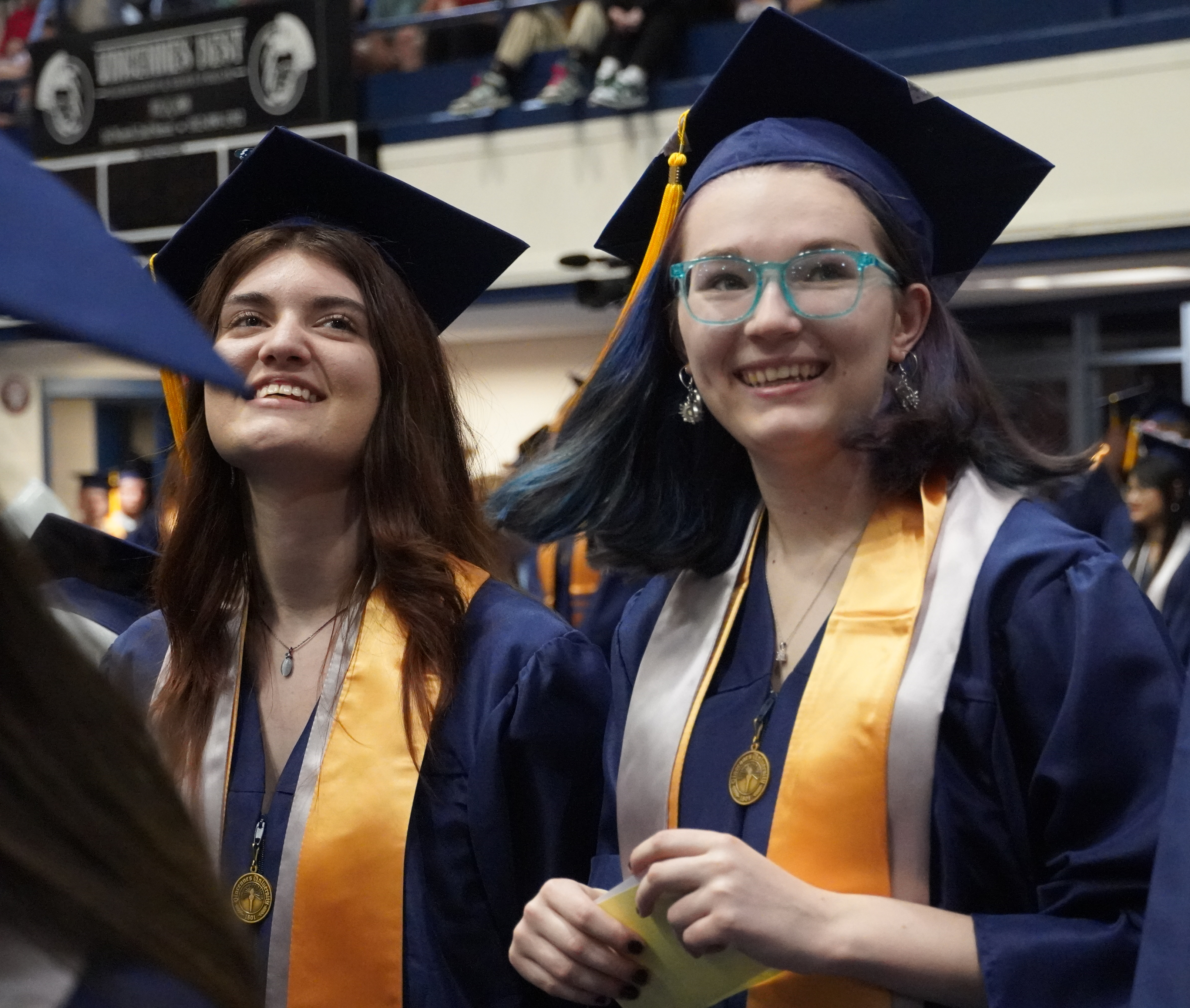 2 smiling female VU graduates wearing caps and gown. One is wearing glasses and has brightly colored hair.